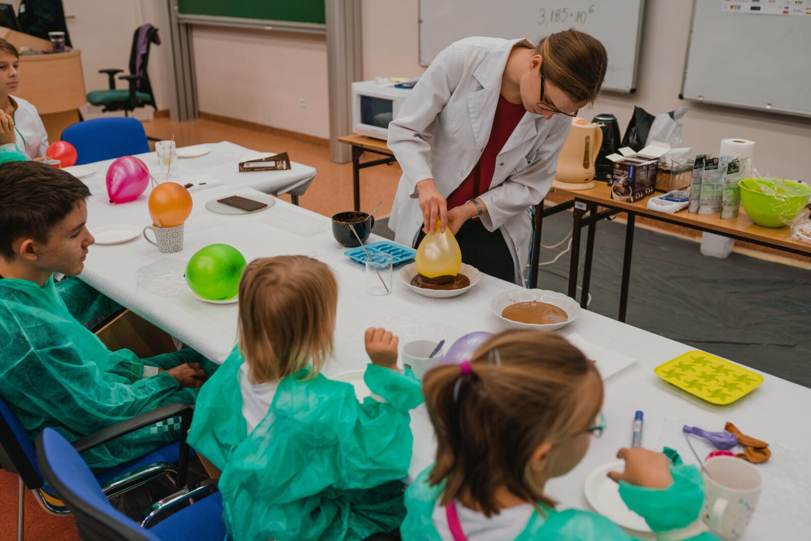 młoda kobieta w białym rozpiętym fartuchu, w okularach trzyma żółty balon, który macza w brązowym płynie, z przodu dzieci siedzące przy stole, ubrane w zielone fartuchy, na stole balony w naczyniach.