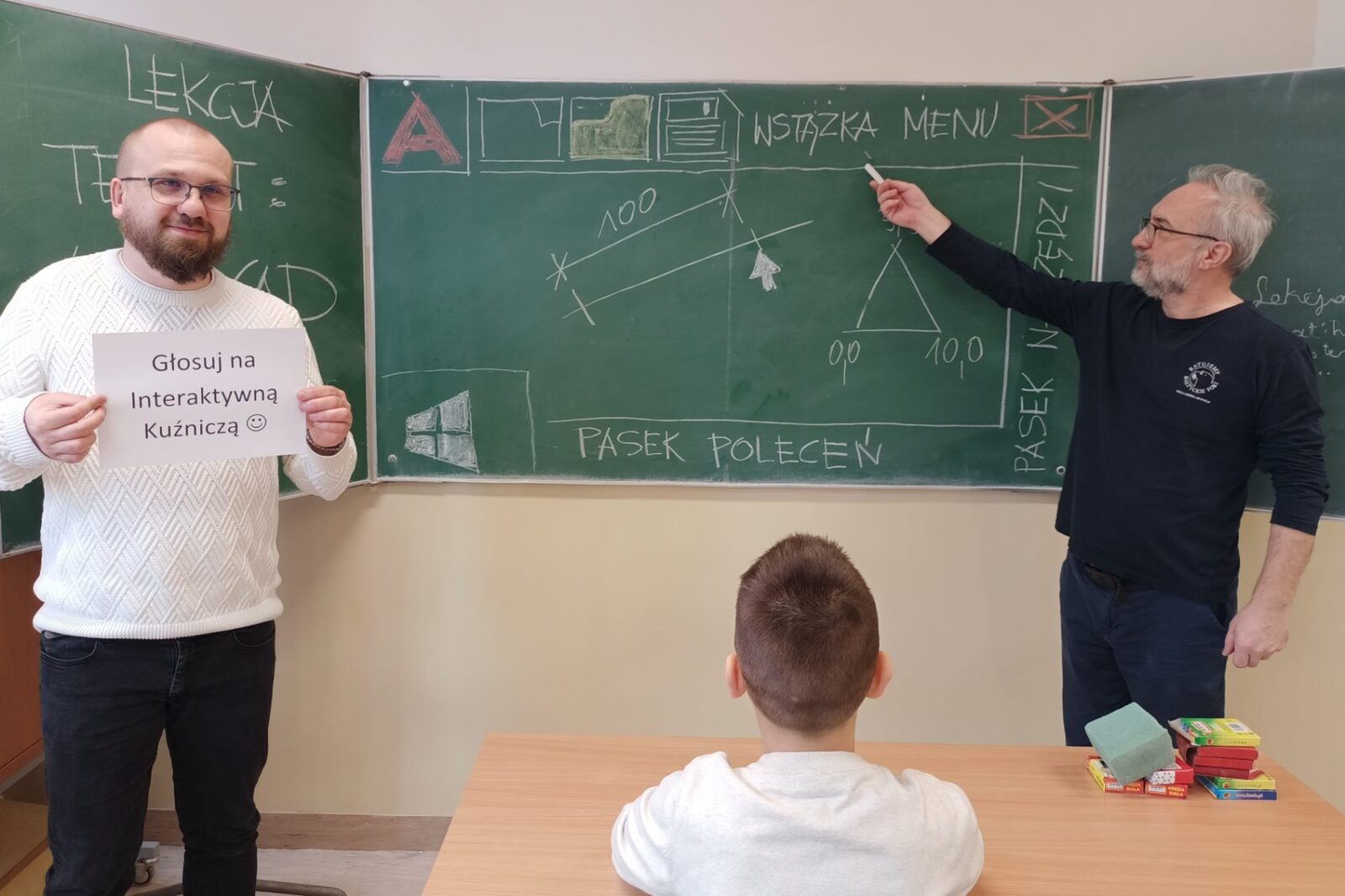 Dwóch mężczyzn stoi przy tablicy, jeden wskazuje na tablicę kredą, a drugi trzyma kartkę z napisem: Głosuj na Interaktywną Kuźniczą
