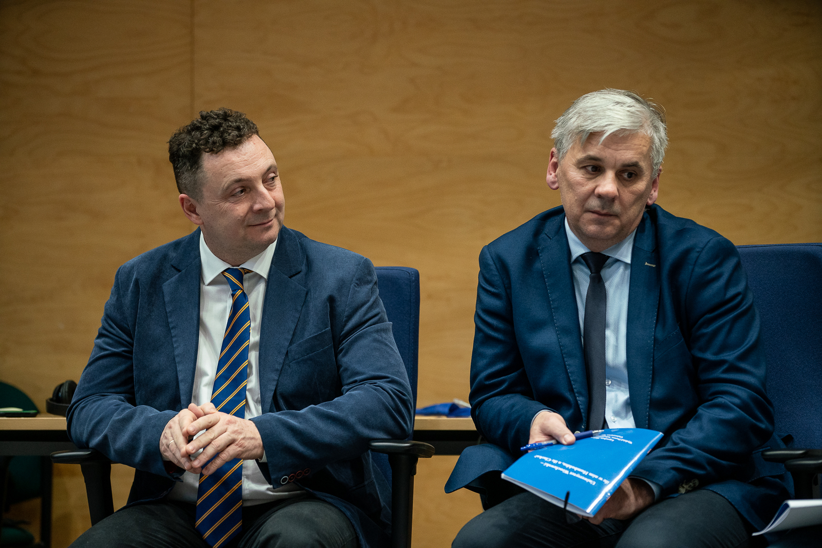 dwaj meżczyżni w średnim wieku w marynarkach siedzący na krzesłach