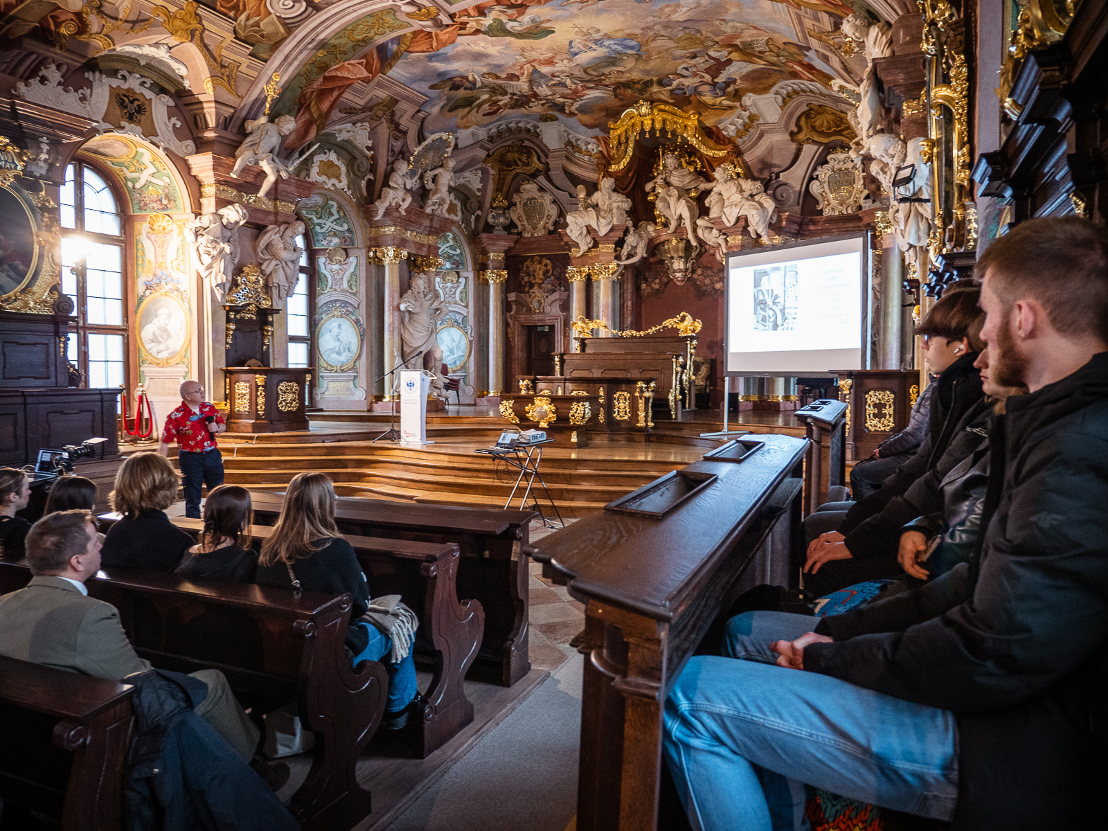zabytkowa historyczna aula, na środku mężczyzna z mikrofonem mówiący do młodych ludzi siedzących w ławach