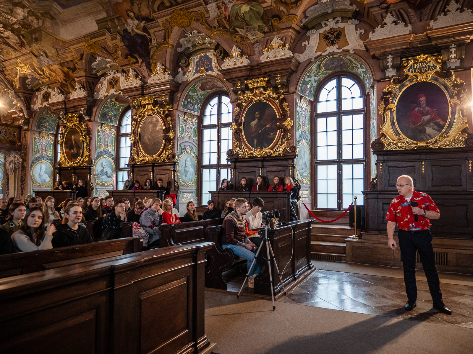 zabytkowa aula ludzie siedzący w ławach , na środku mężczyzna w czerwonej koszuli z mikrofonem