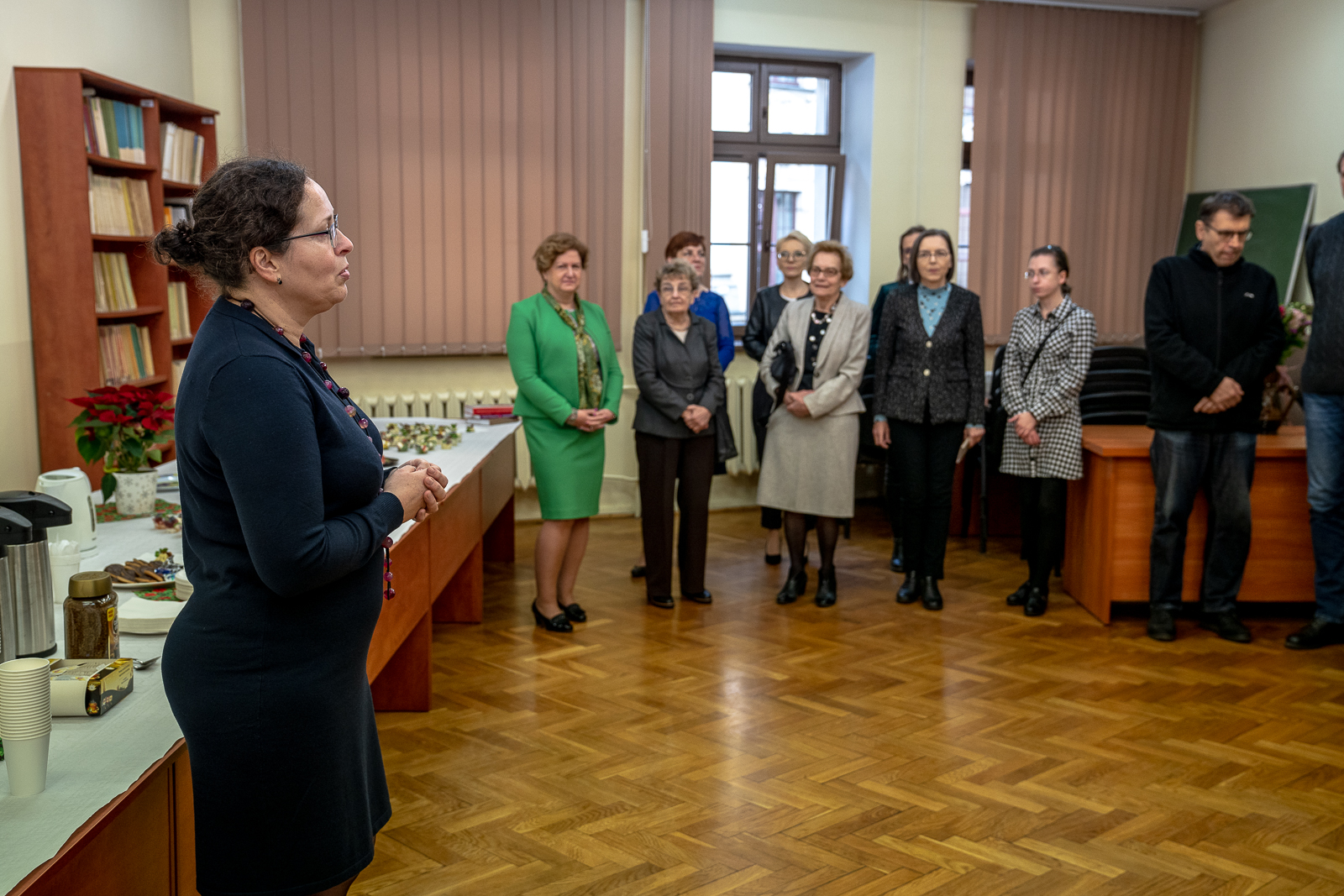 kobieta w granatowej sukience i koralach o brązowych kręconych włosach z okularami, z tyłu grupa kobiet i mężczyzna