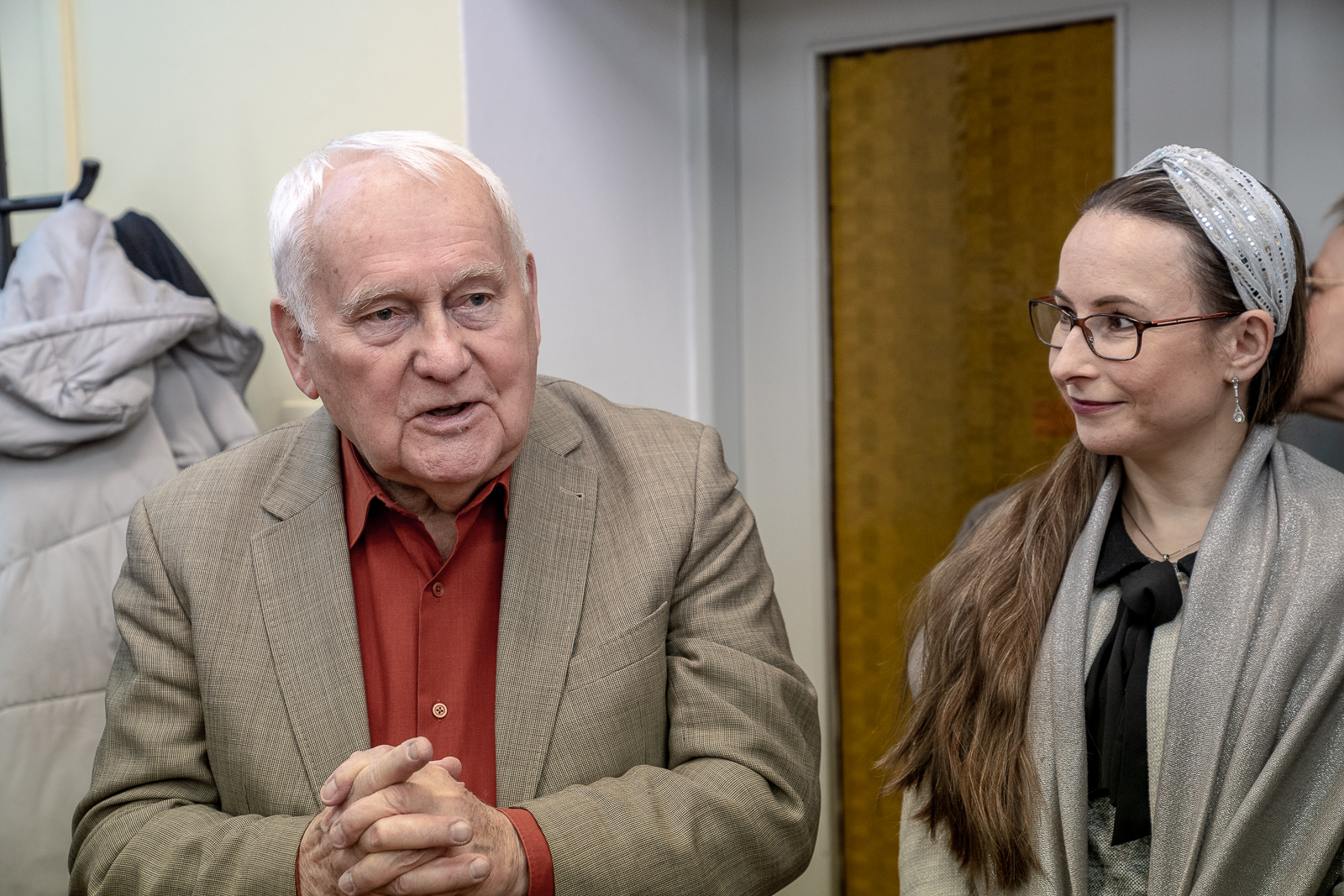 starszy siwy mężczyzna w pomarańczowej koszuli i jasnej marynarce, młoda kobieta w okularach i opasce na głowie