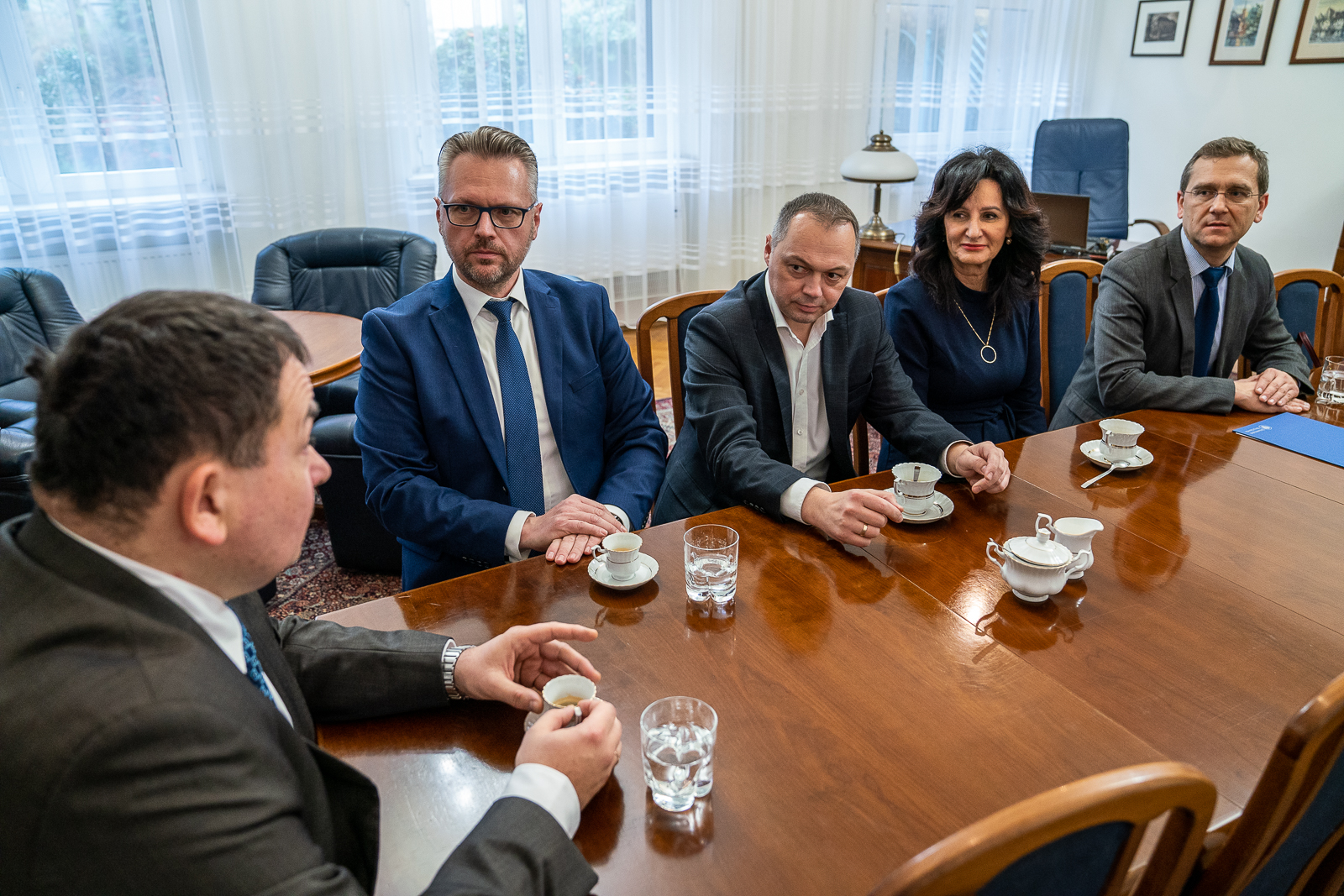Pięć osób ubranych elegancko siedzi przy stole. Na stole filiżanki i szklanki z wodą.