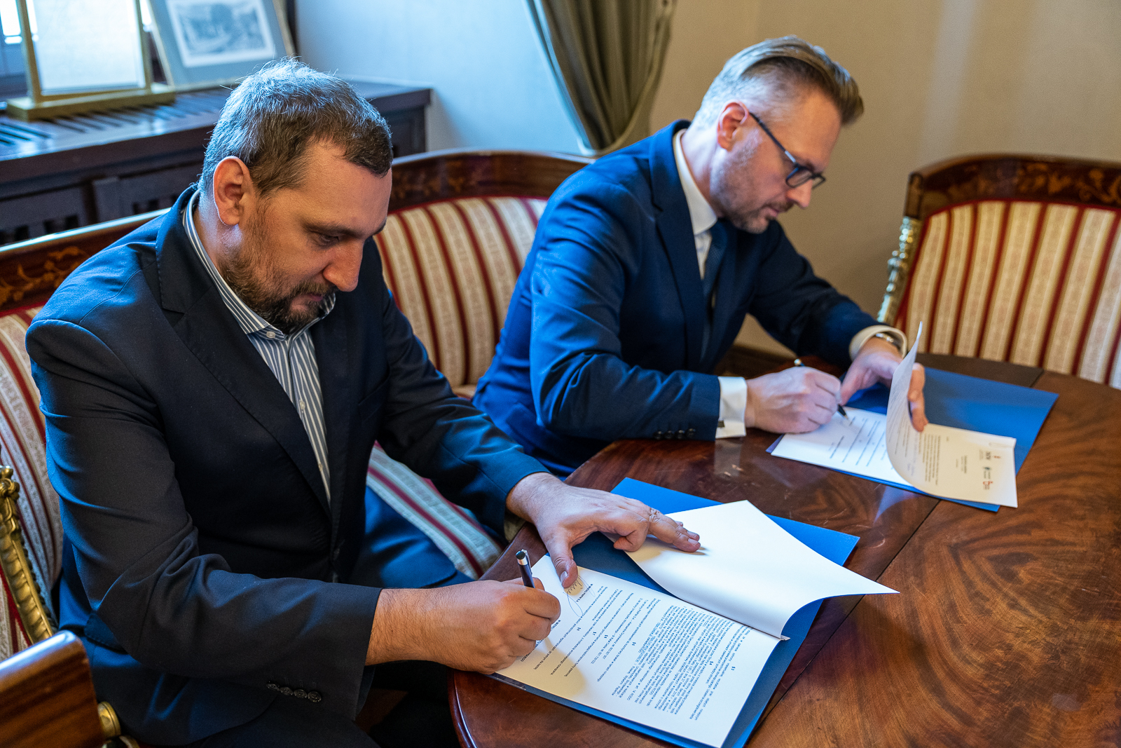 Dwóch panów w garniturach siedzi przy stole i podpisuje dokumenty