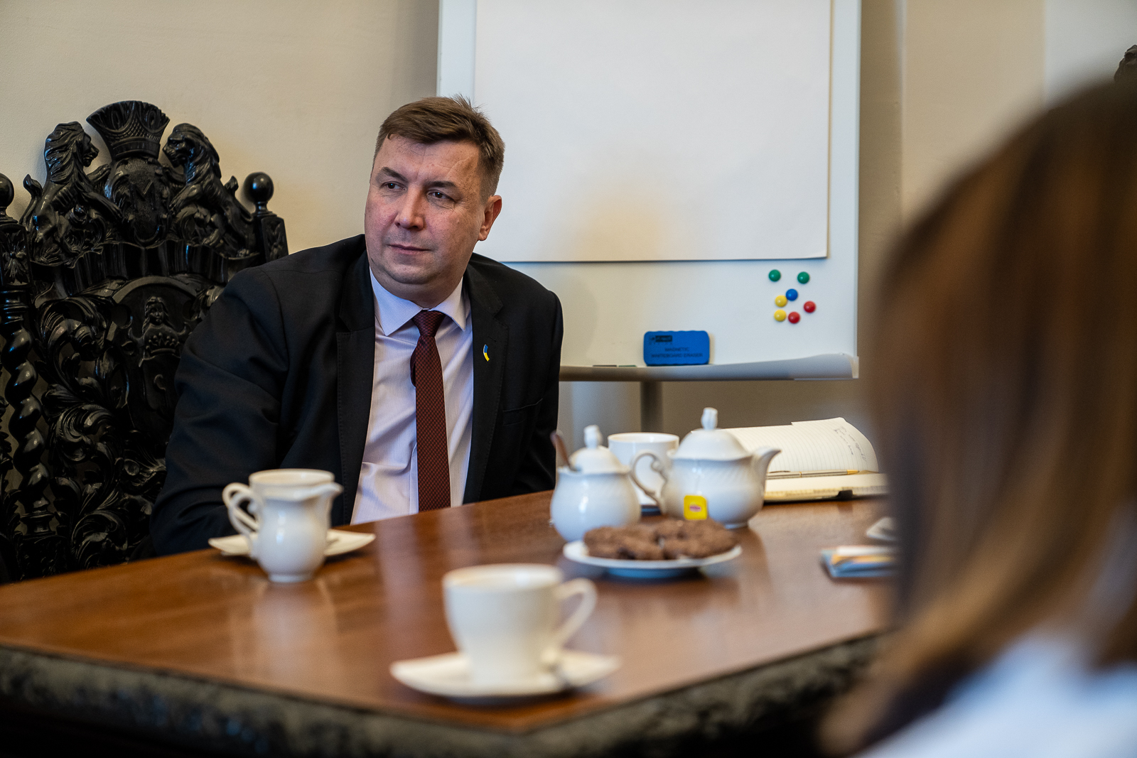 Mężczyzna w garniturze przy stole, na stole filiżanki, za nim biała tablica