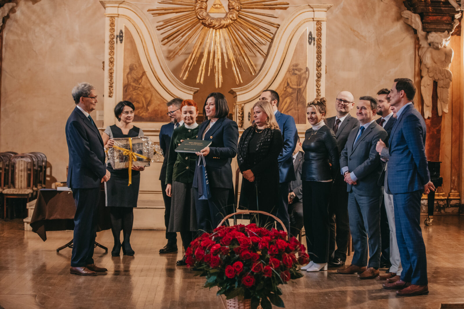 starszy mężczyzna stoi naprzeciw grupy osób, kobieta wręcza mu dużą księgę