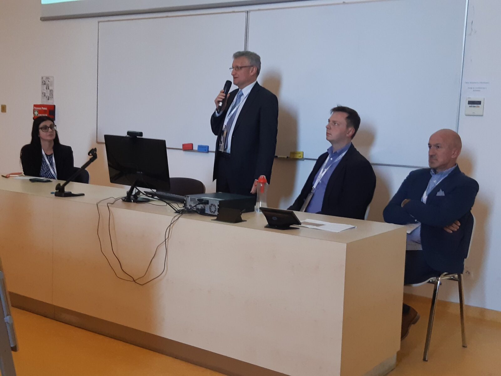 Mężczyzna w gariturze z mikrofonem w ręku stoi za biurkiem na tle białej tablicy, obok niego siedzą dwaj mężczyźni w garniturach