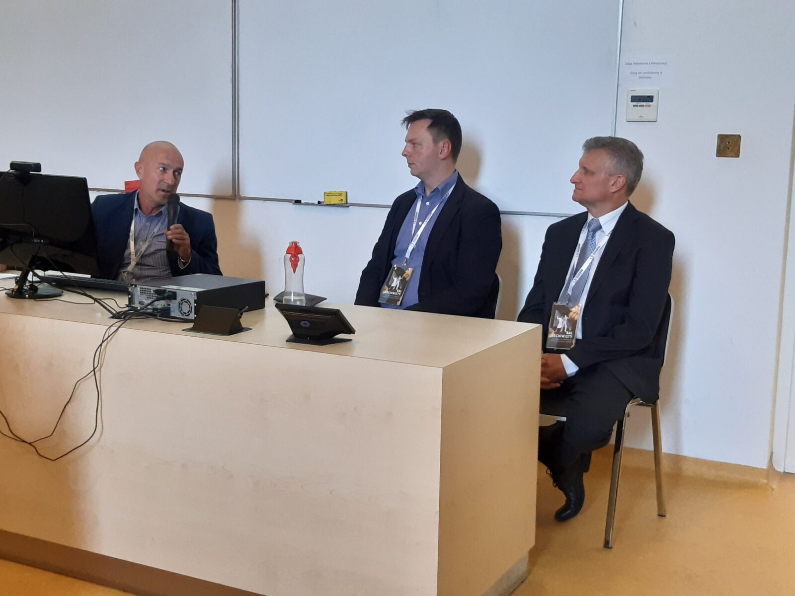 Trzej mężczyźni w garniturach siedzą za biurkiem, za nimi biała tablica