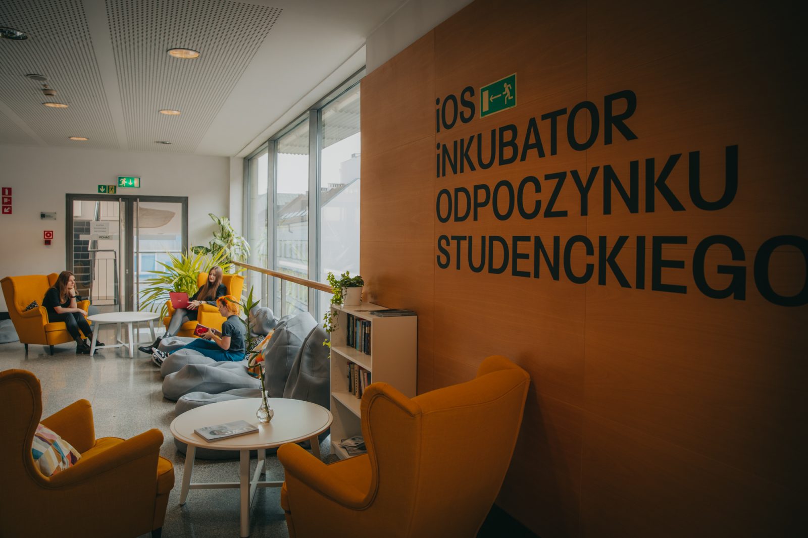 pomieszczenie z dużym oknem, fotelami, stolikami i szafką na książki, napis na ścianie iOS Inkubator Odpoczynku Studenckiego