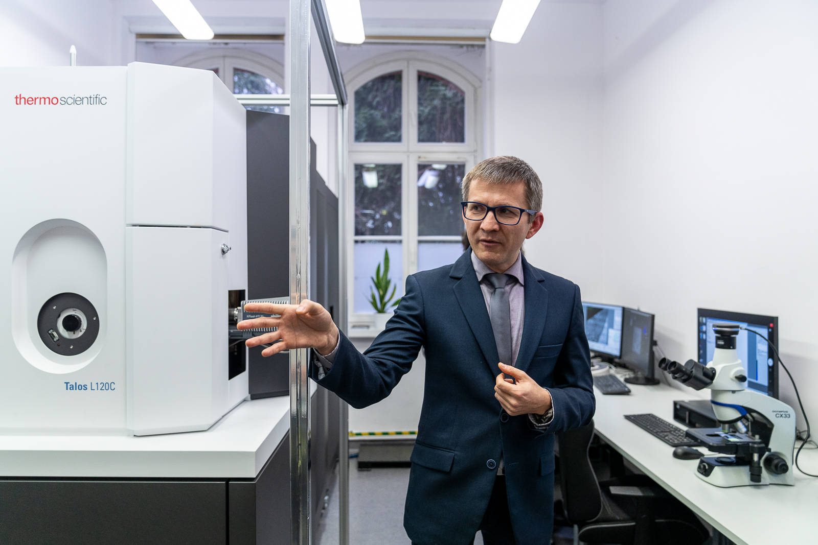 ubrany w garnitur mężczyzna w średnim wieku stojący pomiędzy dużym białym urzedzeniam a biurkami z komputerami