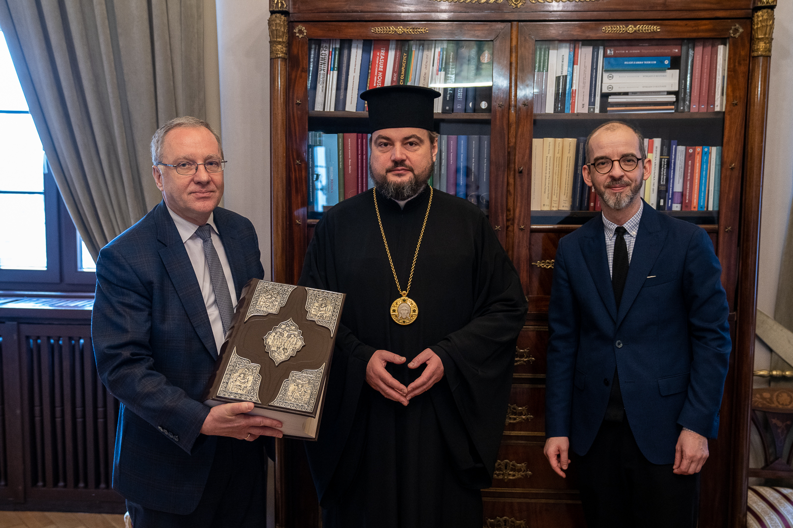 trzej mężczyźni stojący przed szafką z książkami, dwaj stojący bokach w garniturach, w środku ubrany w czarny strój i czarne nakrycie głowy