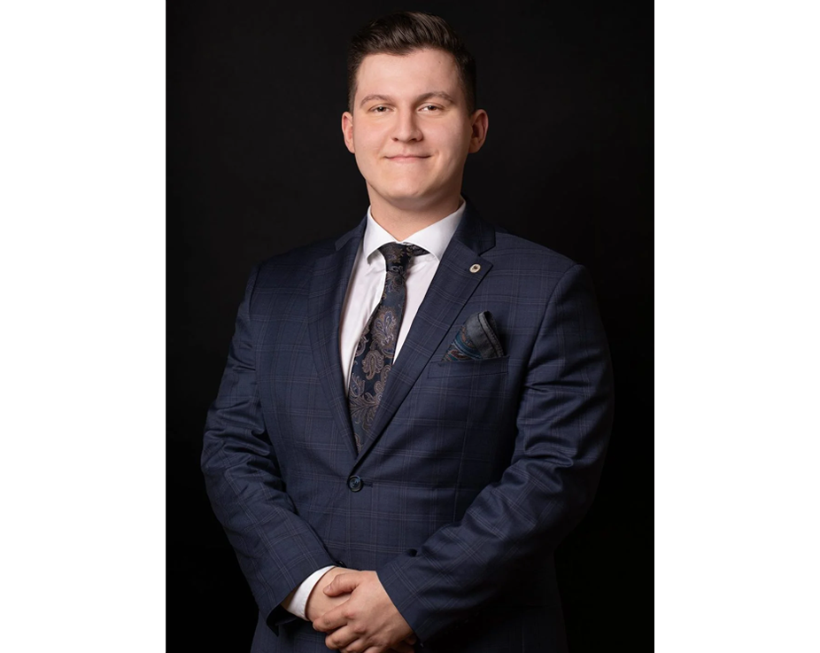 młody mężczyzna w marynarce, białej koszuli z krawatem, splecione dłonie trzyma na wysokości pasa