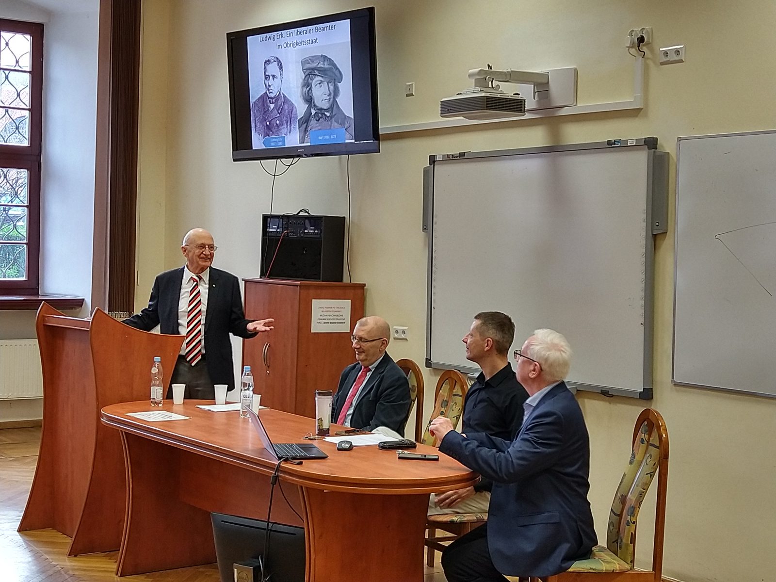 czterech mężczyzn, trzech siedzi przy stole, jeden stoi