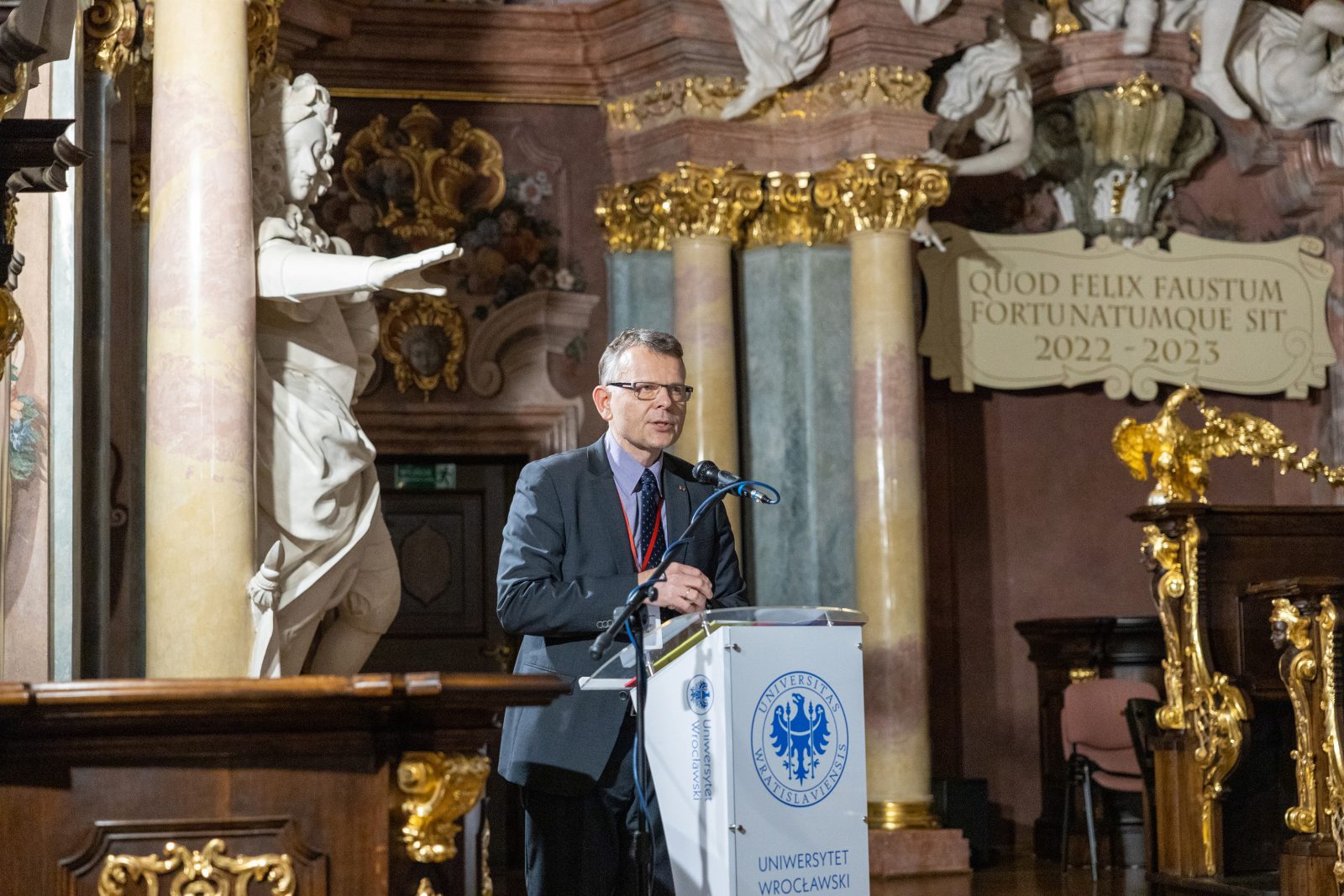 Mężczyzna w garniturze i okularach stoi za mównica w barokowym wnętrzu