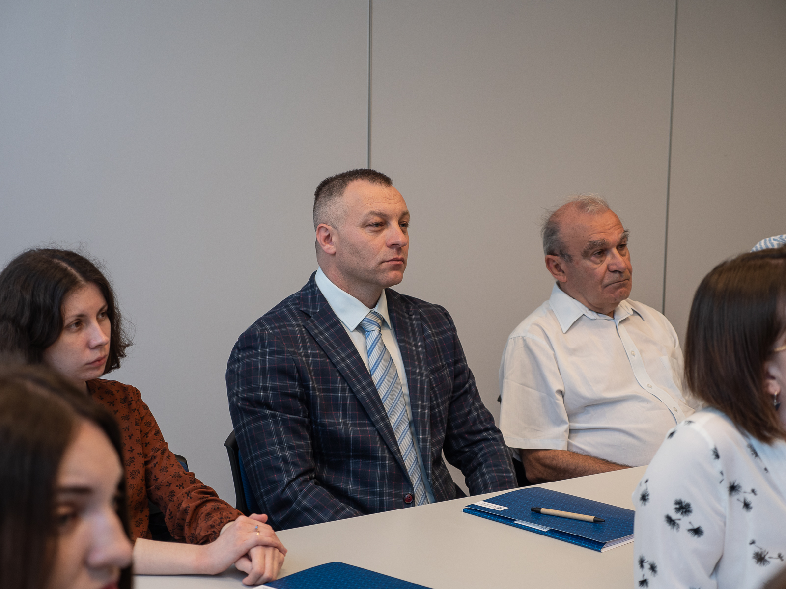Mężczyzna w średnim wieku w garniturze i krawacie siedzący przy stole. Po jego lewej stronie, a prawej na zdjęciu starszy mężczyzna w białej koszuli z krótkim rękawem. Po jego prawej stronie kobieta. Każdy z nich patrzy przed siebie.