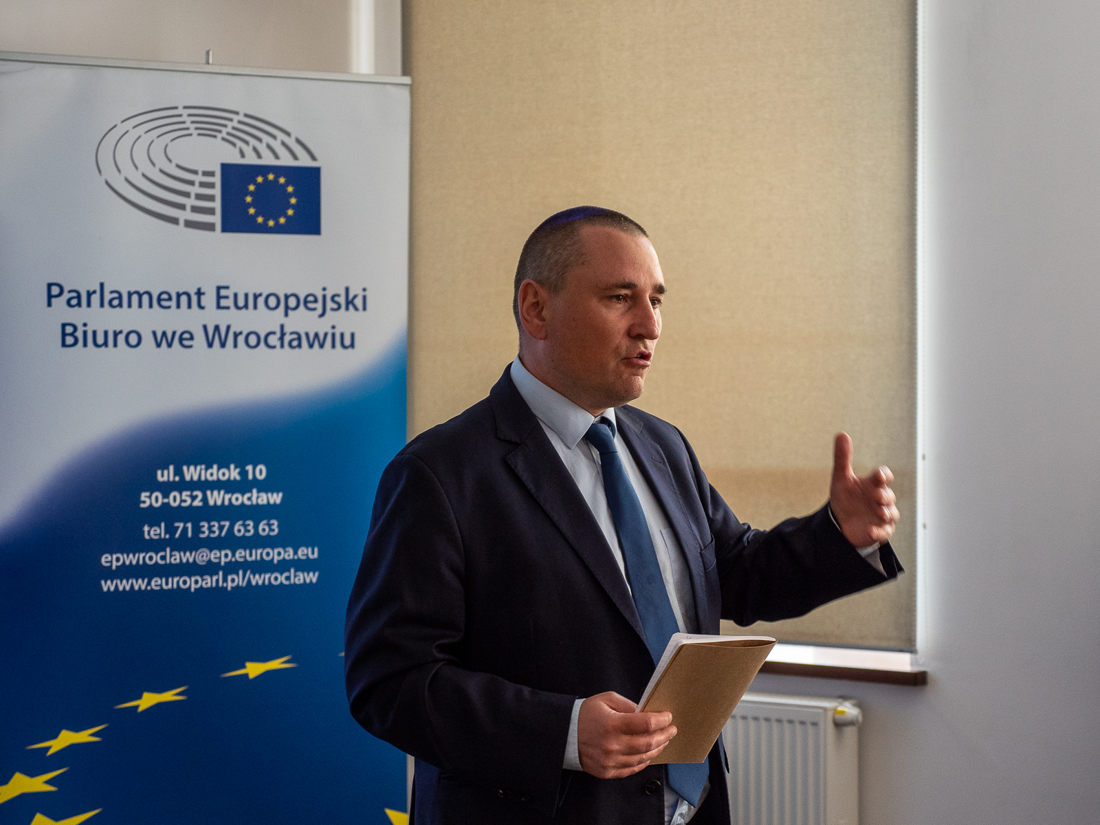 Mężczyzna w garniturze przemawia, gestykuluje jedną ręką, w drugiej trzyma zeszyt. Za jego plecami rollup z napisem "Parlament Europejski Biuro we Wrocławiu" i danymi teleadresowymi