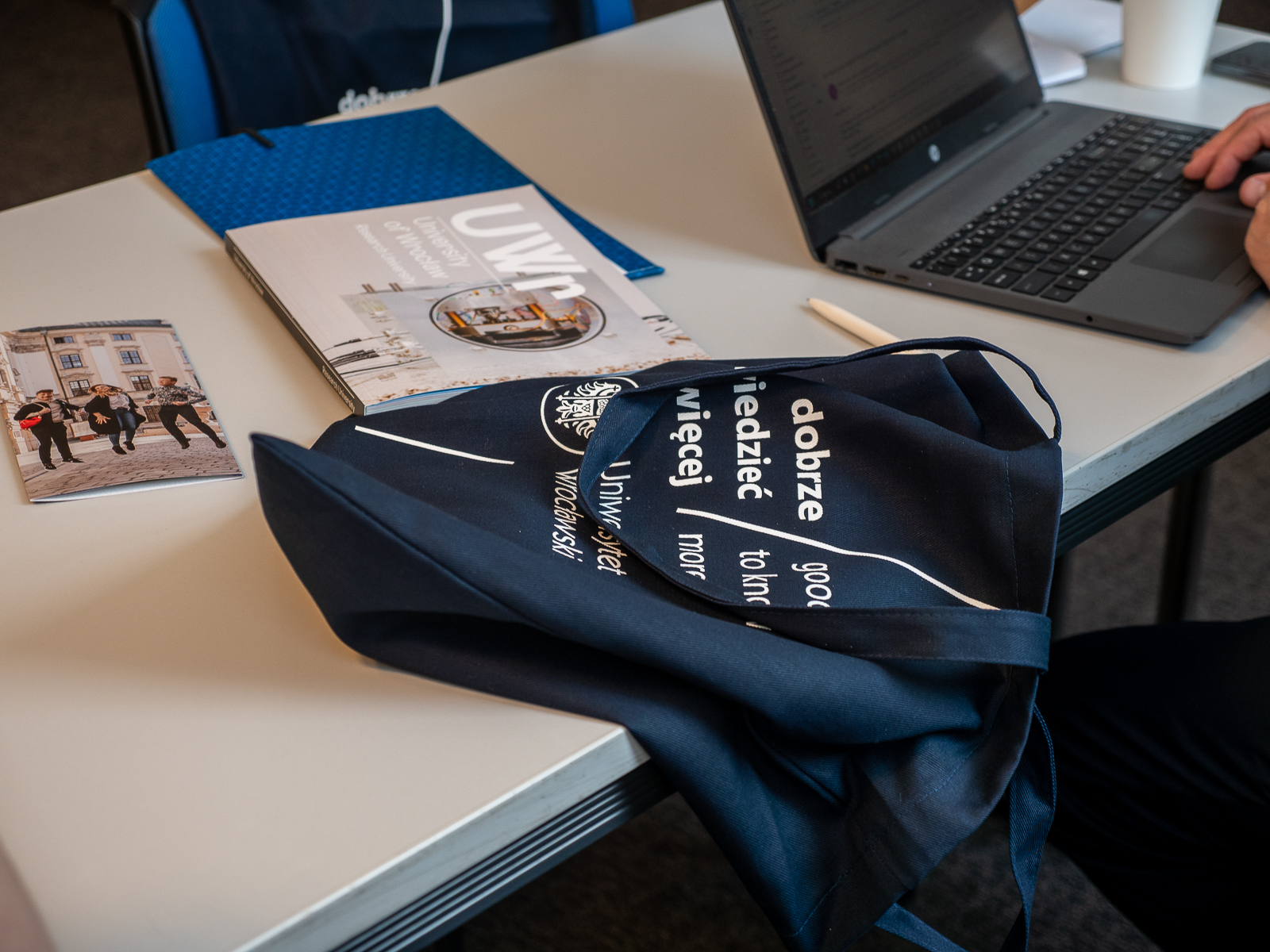 Worek z logo Uniersytetu Wrocławskiego lezący na stole. Obok niego książka i ulotka. Widać też laptop.