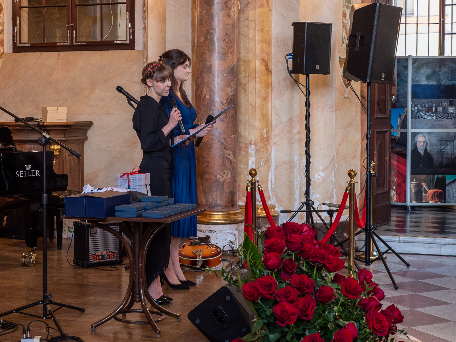 Dwie młode kobiety stojące w zabytkowej sali na podium. Jedna mówi do mikrofonu. Na pierwszym planie bukiet czerwonych róż.