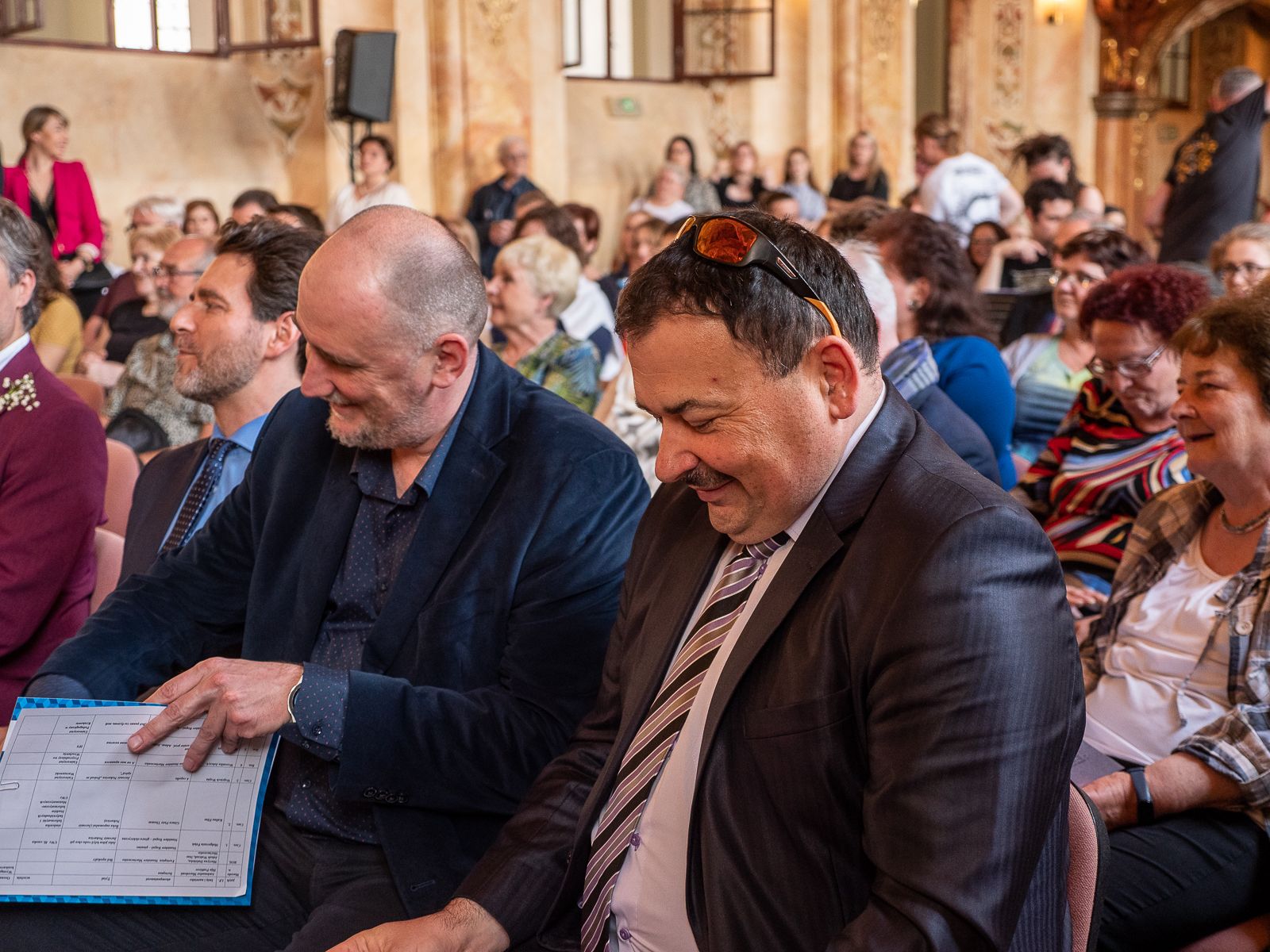 Dwóch uśmiechniętych mężczyzn w ciemnych garinturach. Siedzą w pierwszym rzędzie w zabytkowej sali, za nimi ludzie.