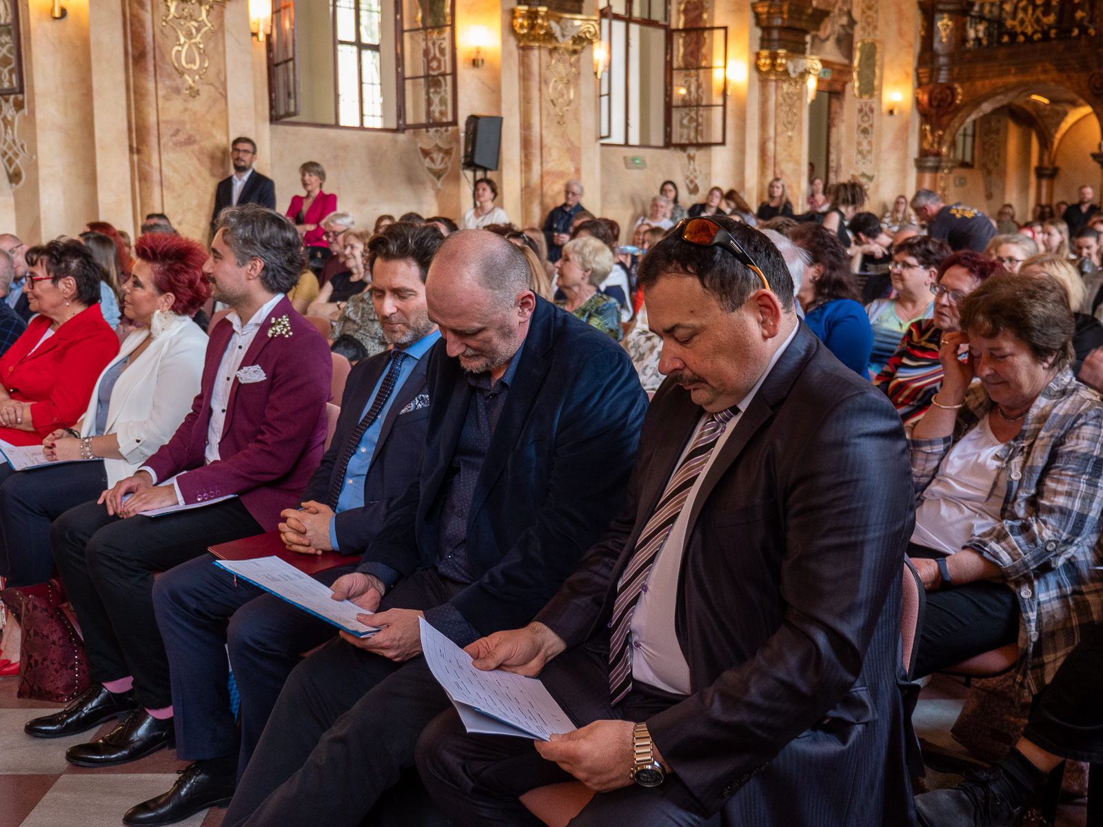 Odświętnie ubrani ludzie w pierwszym rzędzie w zabytkowej sali.