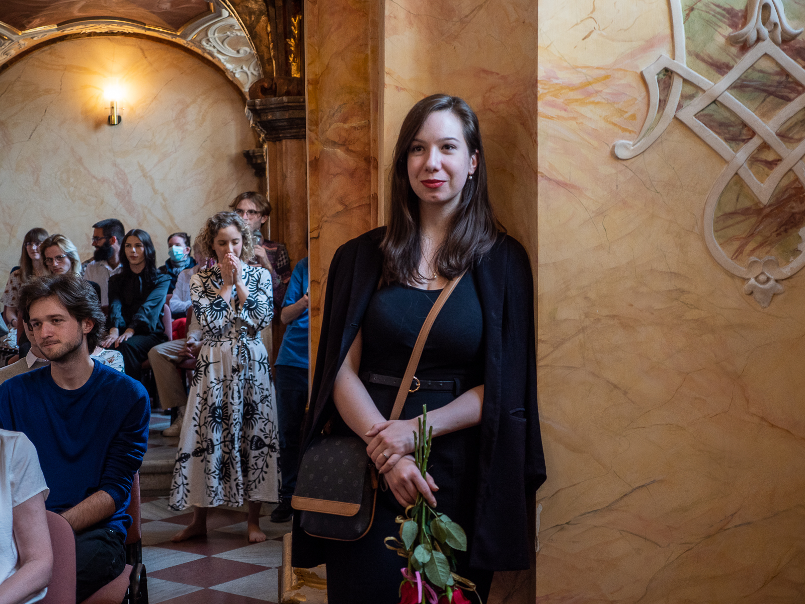 Młoda kobieta w ciemnych włosach, ciemnej marynarnce opiera się o ścianę w zabytkowej sali.