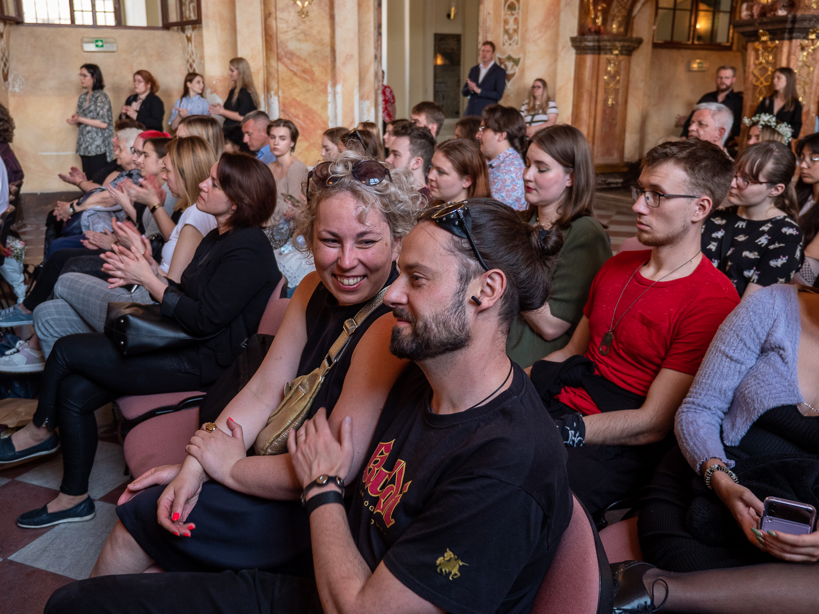 Publiczność zgromadzona w zabytkowej sali. Na pierwszym planie mężczyzna z ciemną brodą i uśmiechnięta kobieta.