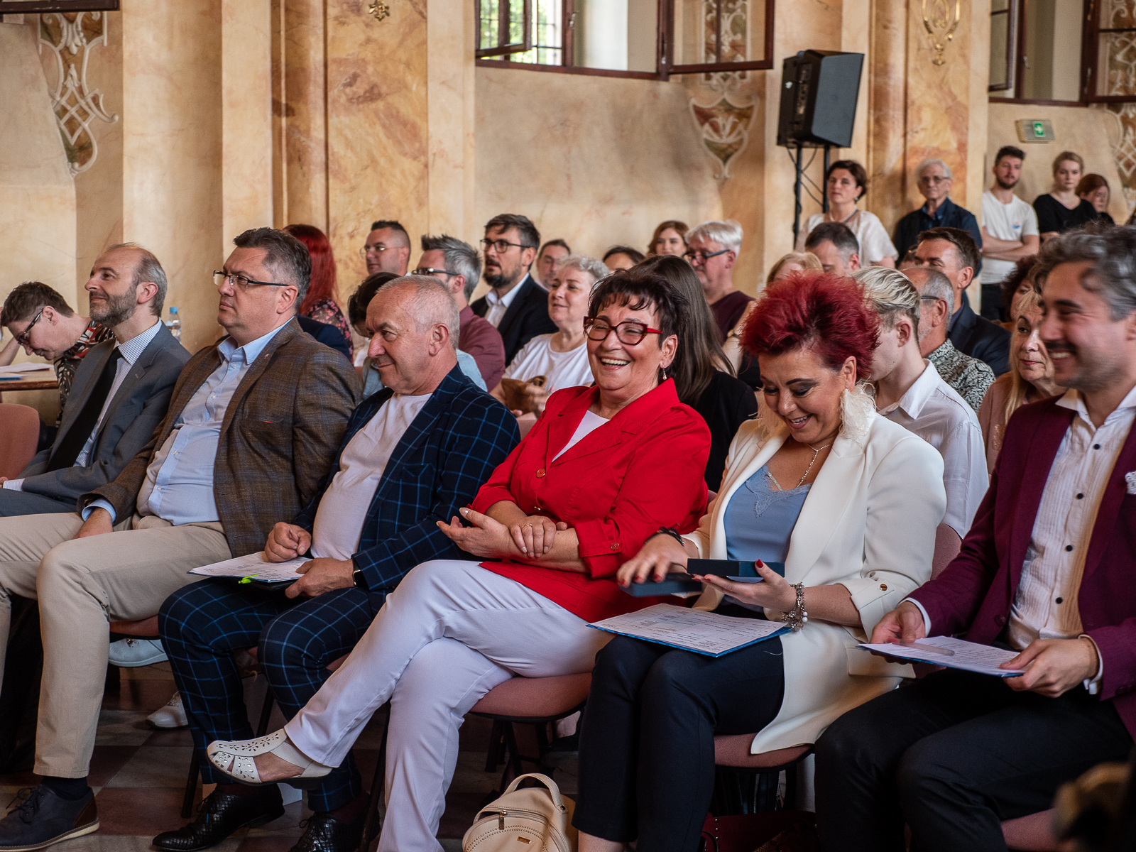Publiczność zgromadzona w zabytkowej sali. W centralnej części zdjęcia kobieta w średnim wieku w cienmnych krótkich włosach, ubrana w czerwoną marynarkę i białe spodnie.