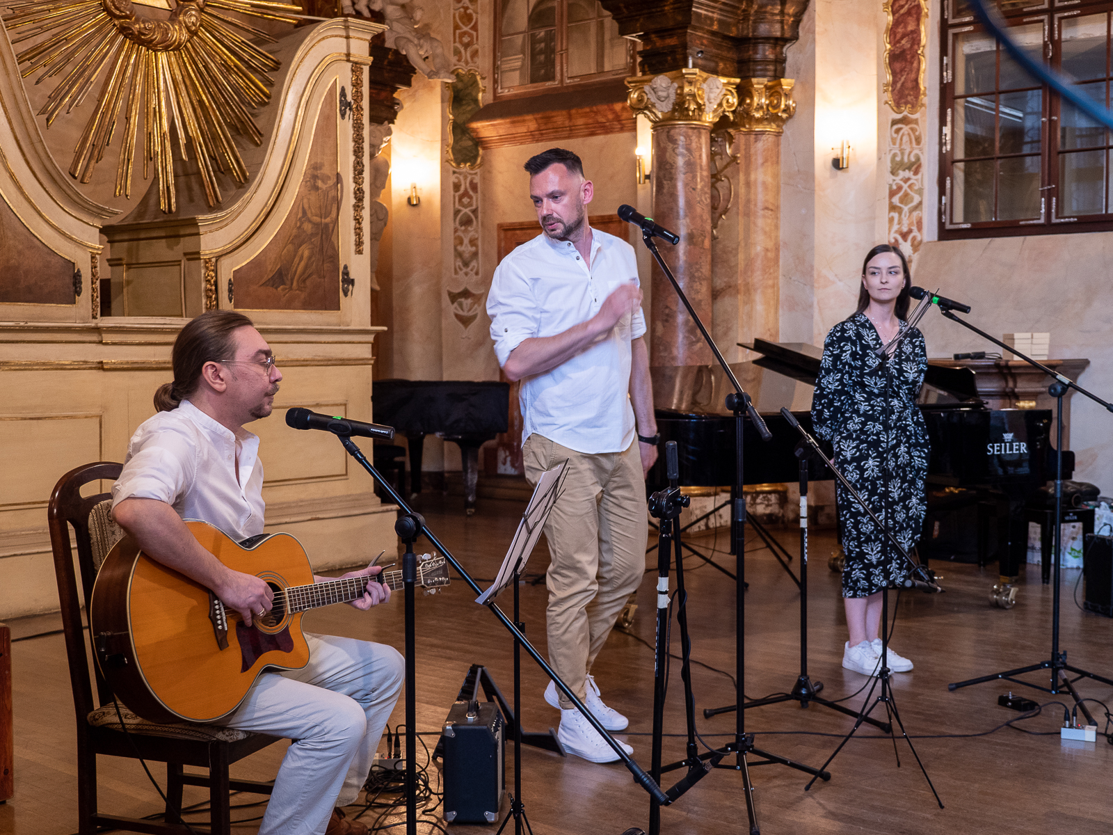Na scenie mężczyzna w białej koszuli grający na gitarze. W środku mężczyzna w białej koszuli i beżowych spodniach w białych butach. Na dalszym planie młoda kobieta w czarnej sukience we wzory.
