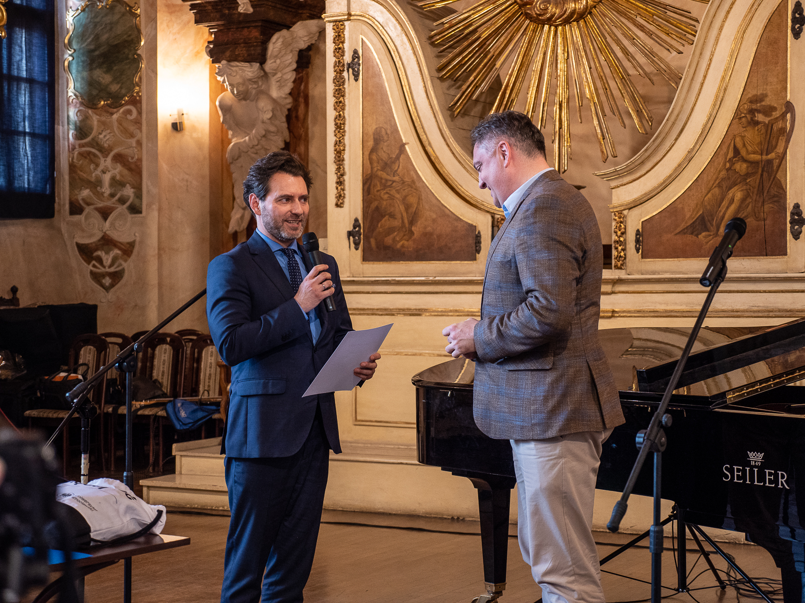 Na scenie dwóch mężczyzn - po lewej stronie młody mężczyzna w ciemnym garniturze z mikrofonem w ręce. Obok niego mężczyzna w ciemnej marynarce.