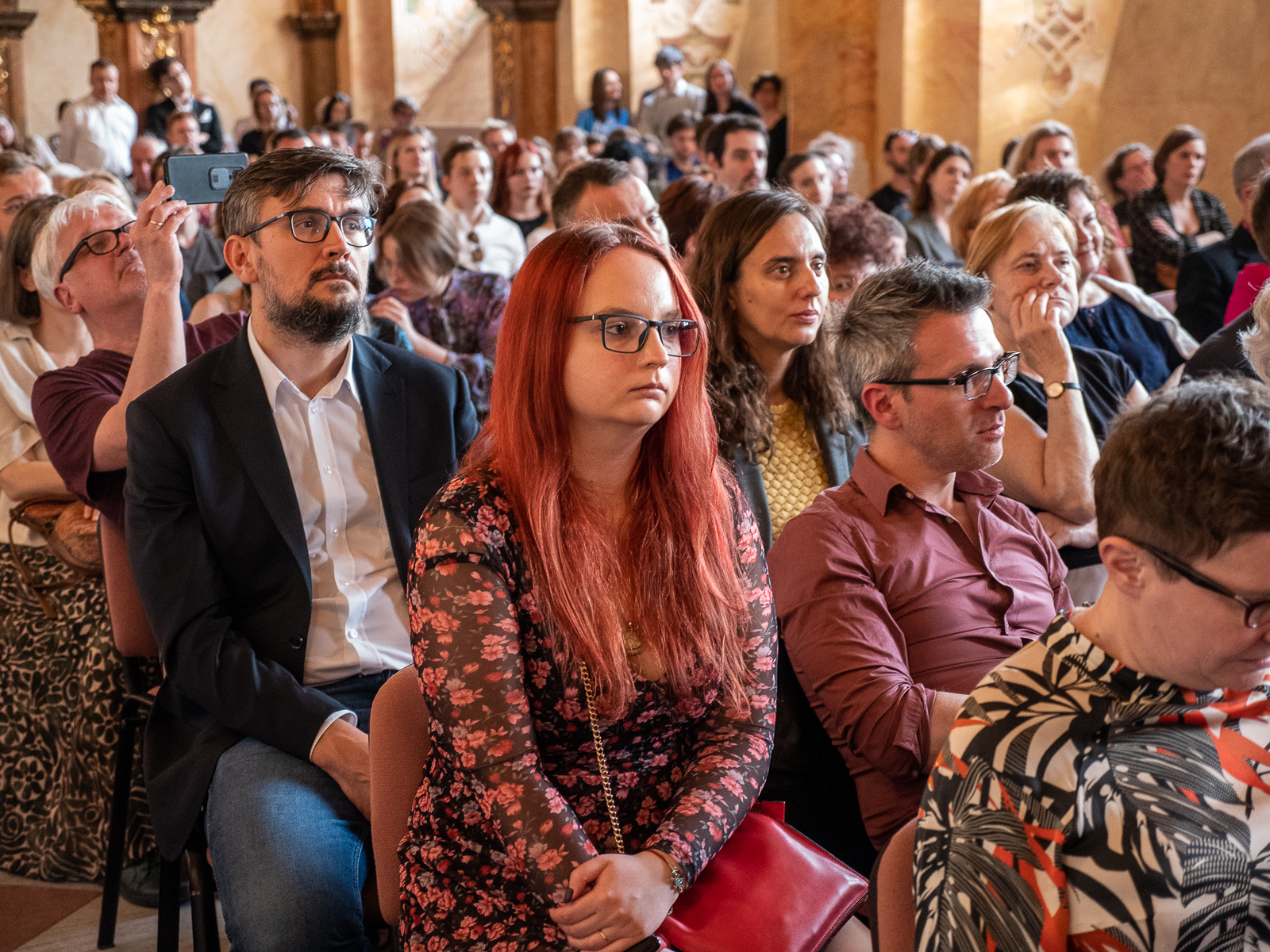 Publiczność w zabytkowej sali. Na pierwszym planie młoda kobieta w rudych włosach i okularach korekcyjnych.