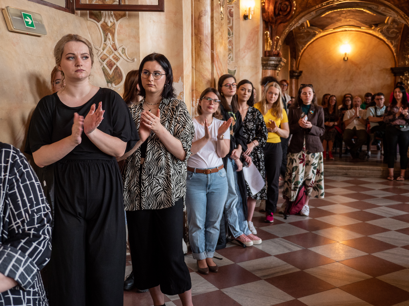Stojąca pod ścianą zabytkowej sali publiczność. Klaszczą w ręce.