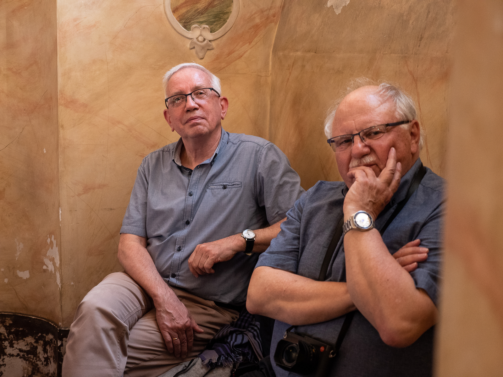 Dwóch starszych mężczyzn z wisymi włosami w niebieskich koszulach siedzący pod ścianą w zabytkowej sali.