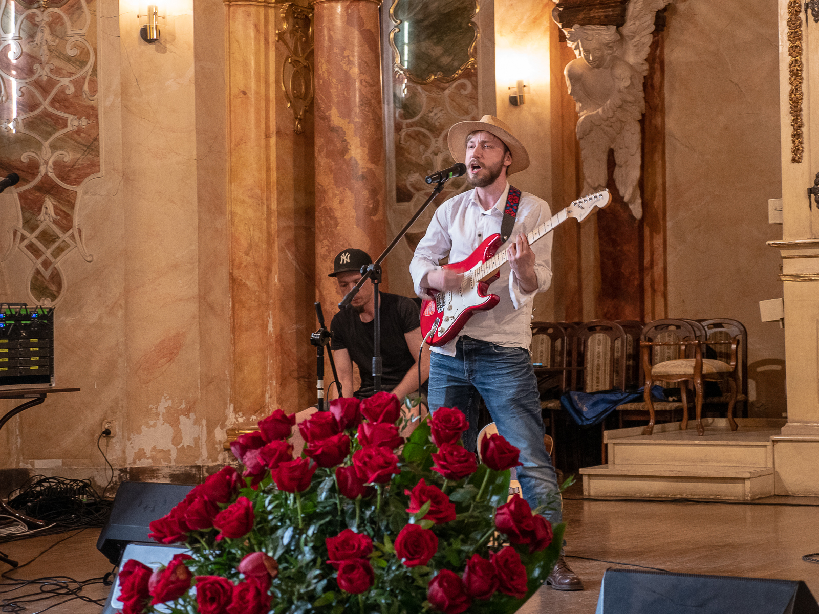 Mężczyzna w białej koszuli i kapeluszu grający na gitarze.