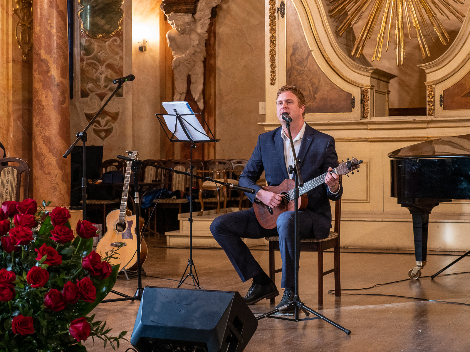 Mężczyzna w ciemnym garniturze siedzi na scenie, gra na gitarze i śpiewa do mikrofonu.
