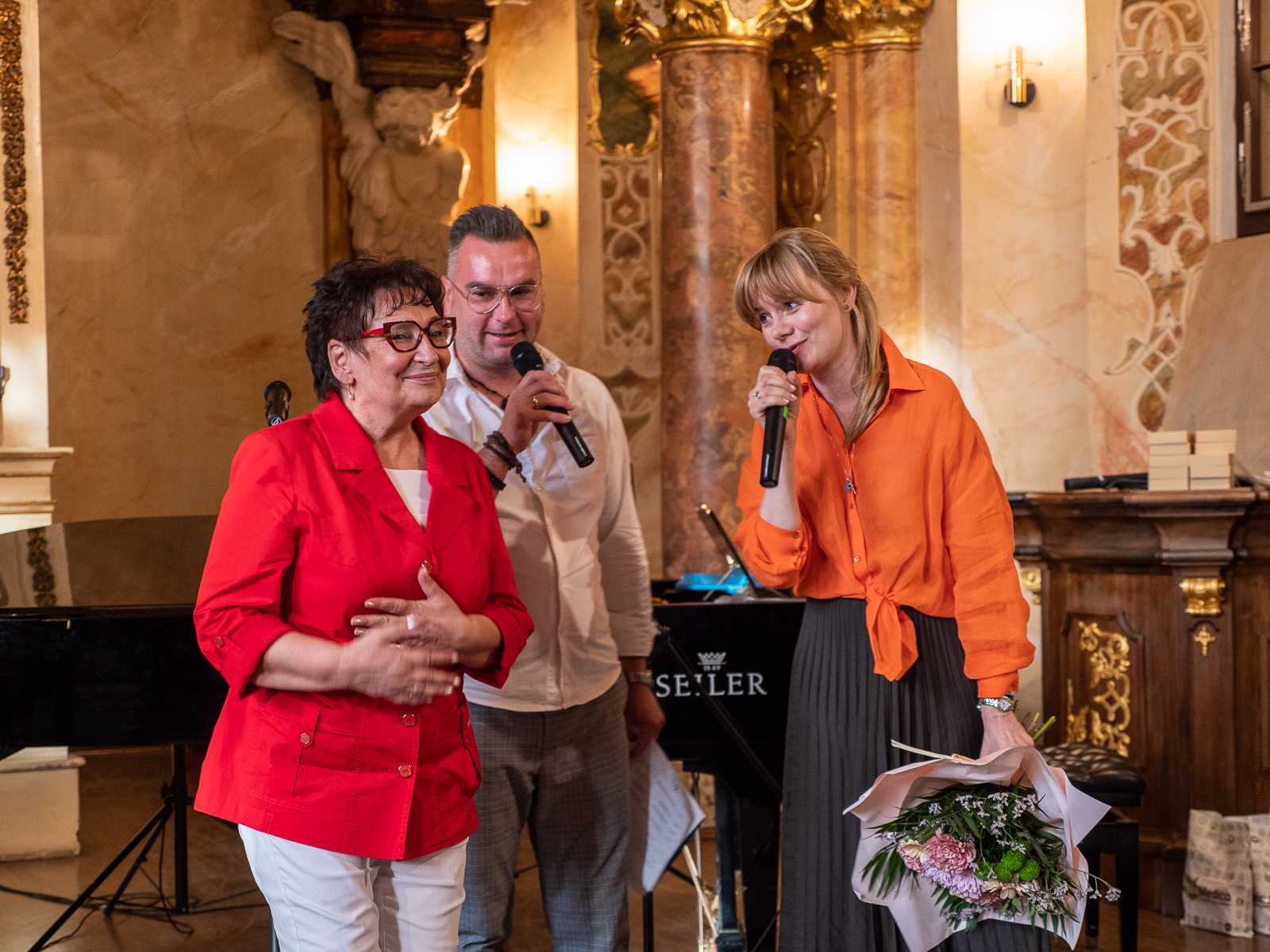 Na scenie kobieta w czerwonej marynarce, mężczyzna w białej koszuli, młoda kobieta w jasnych włosach i pomarańczowej koszuli mówiąca do mikrofonu.