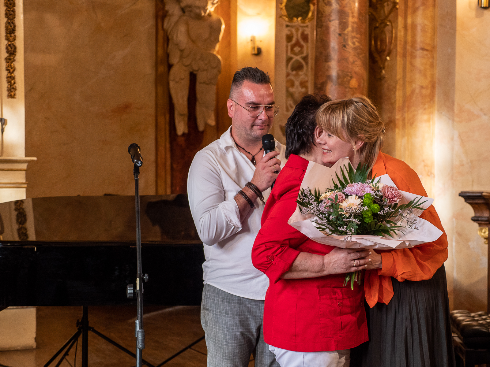 Kobieta w czerwonej marynarce i młoda kobieta w pomarańczowej koszuli przytulają się. Kobieta trzyma bukiet kwiatów. Za nimi mężczyzna w białej koszuli mówiący do mikrofonu.