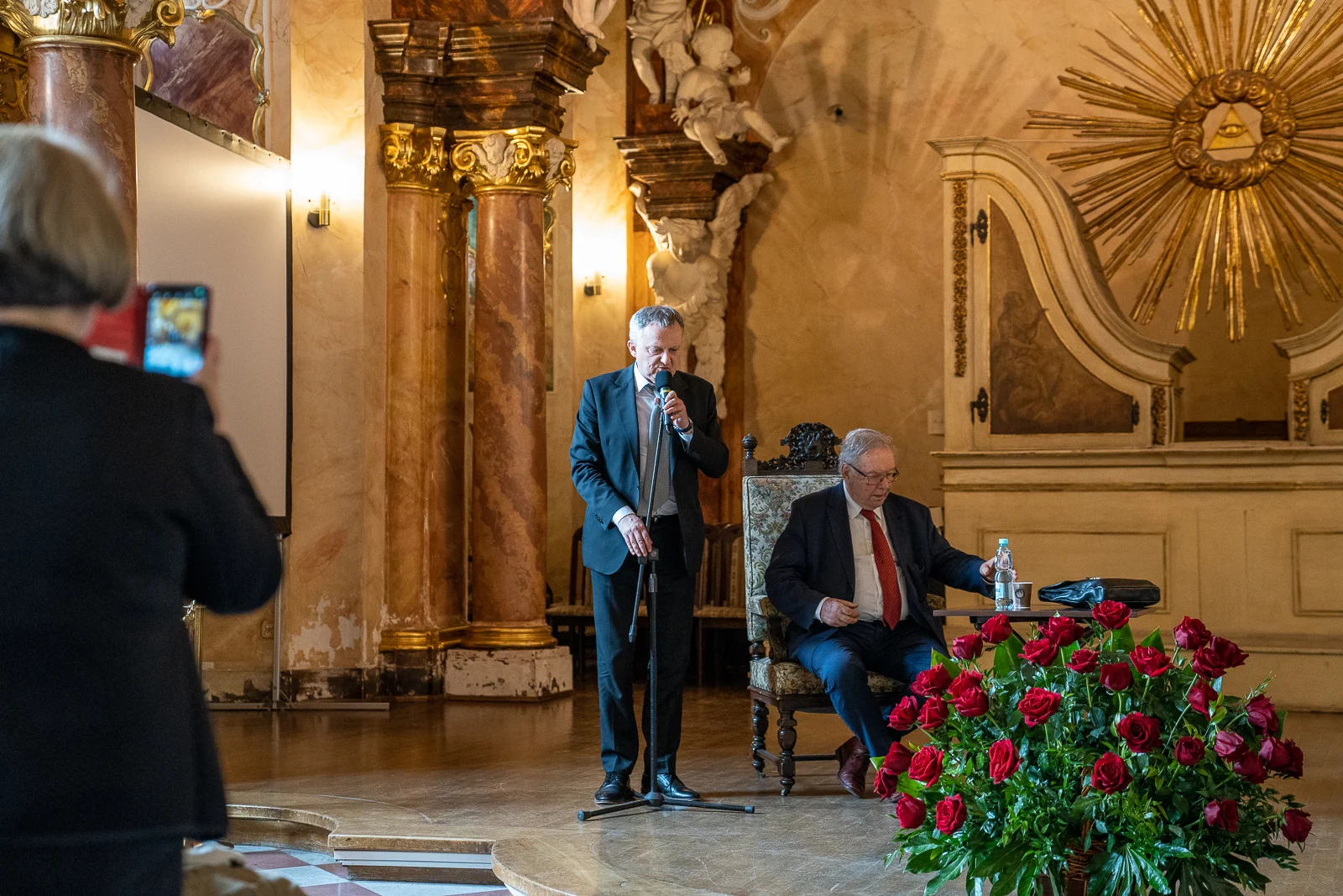 Dwaj meżczyźni w garniturach, po prawej stronie stojący, po lewej stronie siedzący na krześle na tle sali z wieloma zdobieniami, kolumnami i bukietem czewonych róż na pierwszym planie