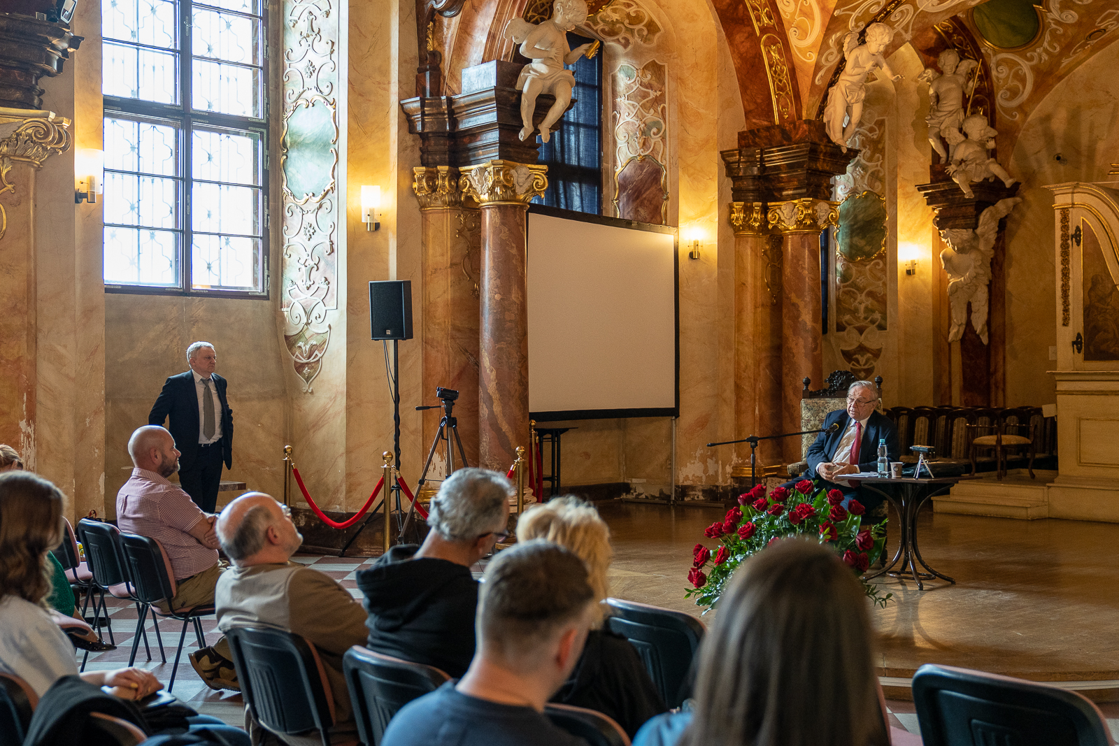 starszy mężczyzna siedzi na krześle przed widzami w sali z wieloma zdobieniami