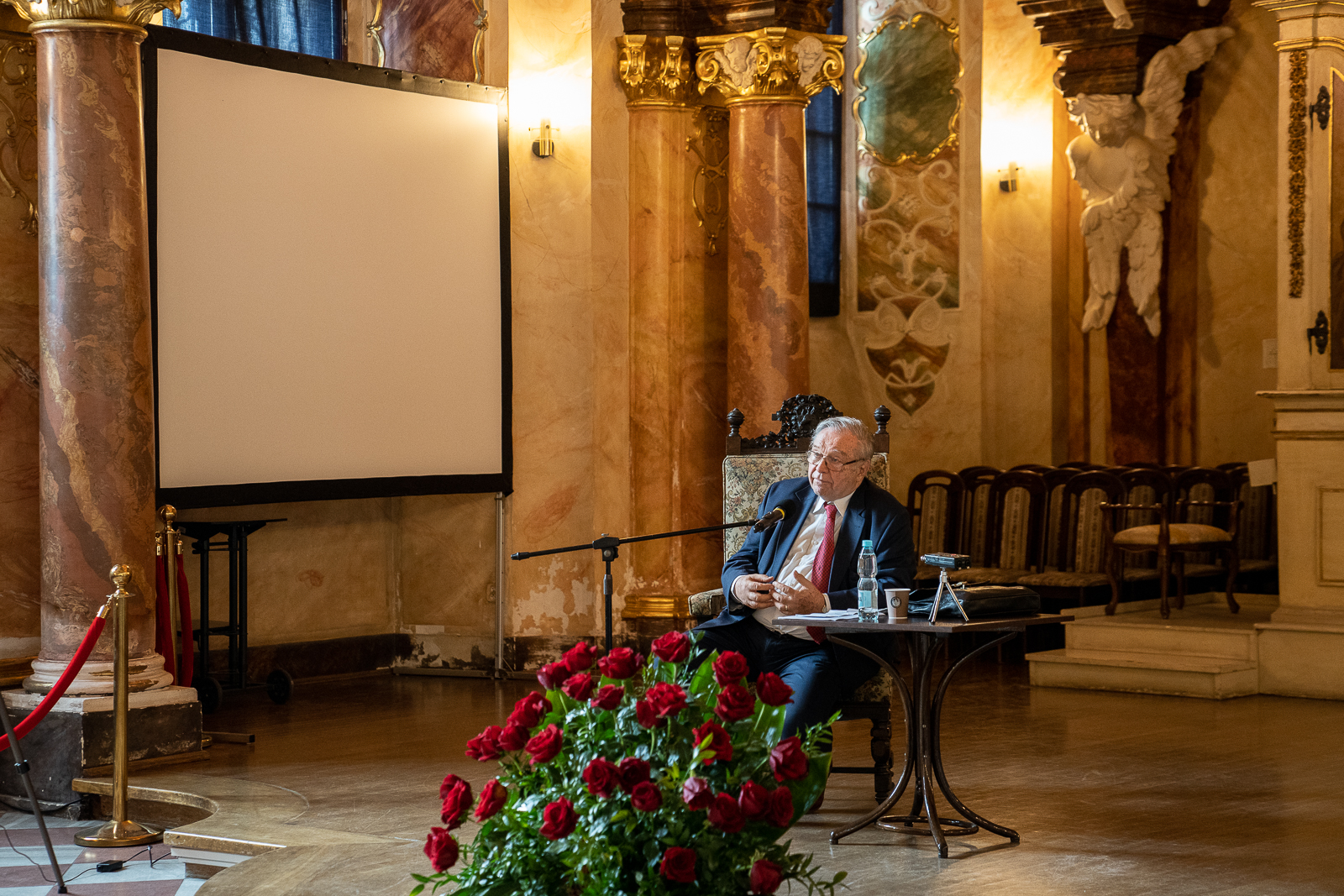 starszy mężczyzna siedzi na krześle przy stoliku i statywie z mikrofonem, przed nim stoi duży bukiet czerwonych róż