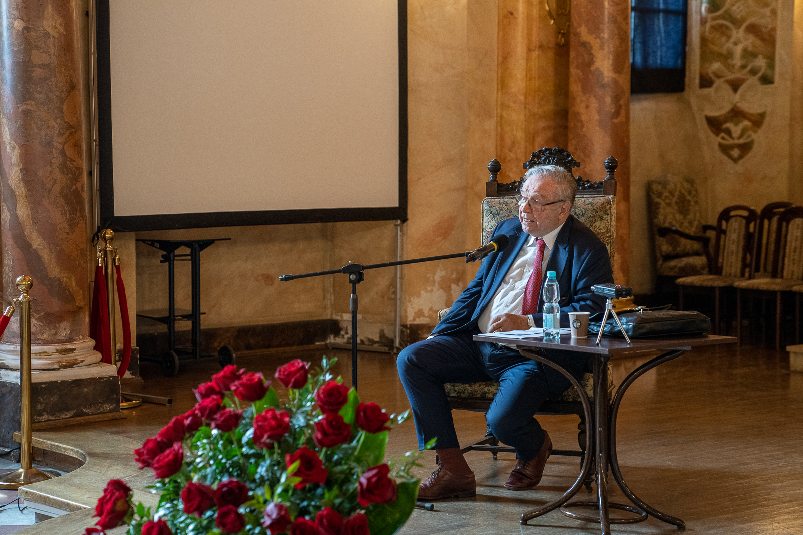 starszy mężczyzna siedzi na krześle przy stoliku i statywie z mikrofonem, przed nim stoi duży bukiet czerwonych róż