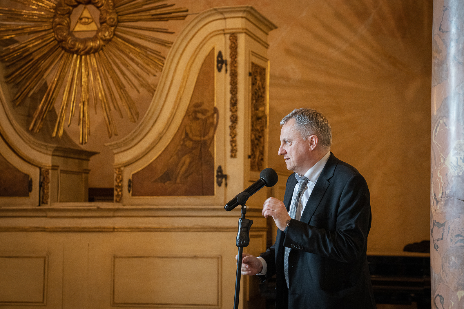 Mężczyzna w garniturze stoi przy statywie z mikrofonem, w tle ozdobne złote elementy wystroju sali