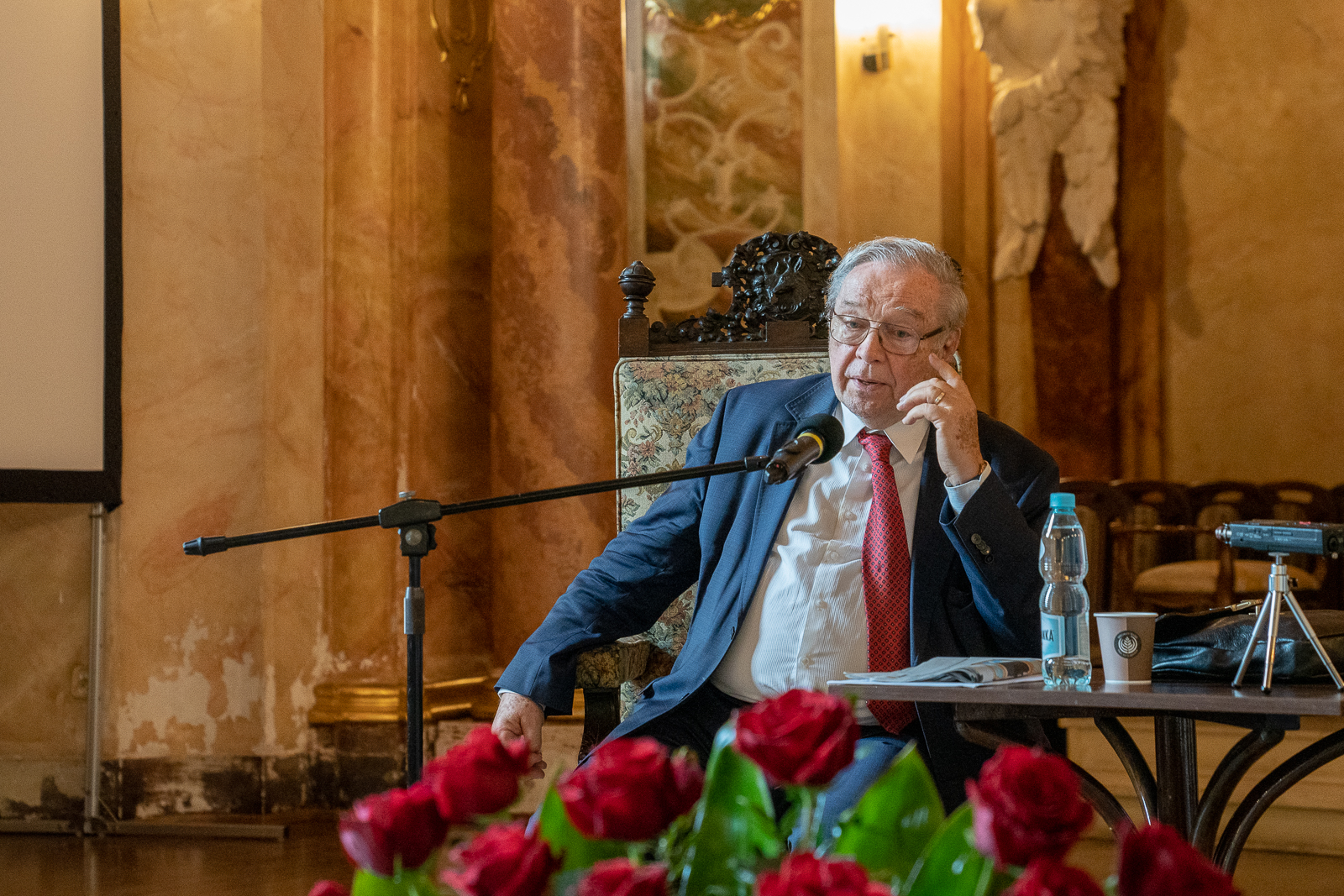 starszy mężczyzna ubrany oficjalnie siedzący na krześle przy stoliku w sali ozdobionej złotymi elementami