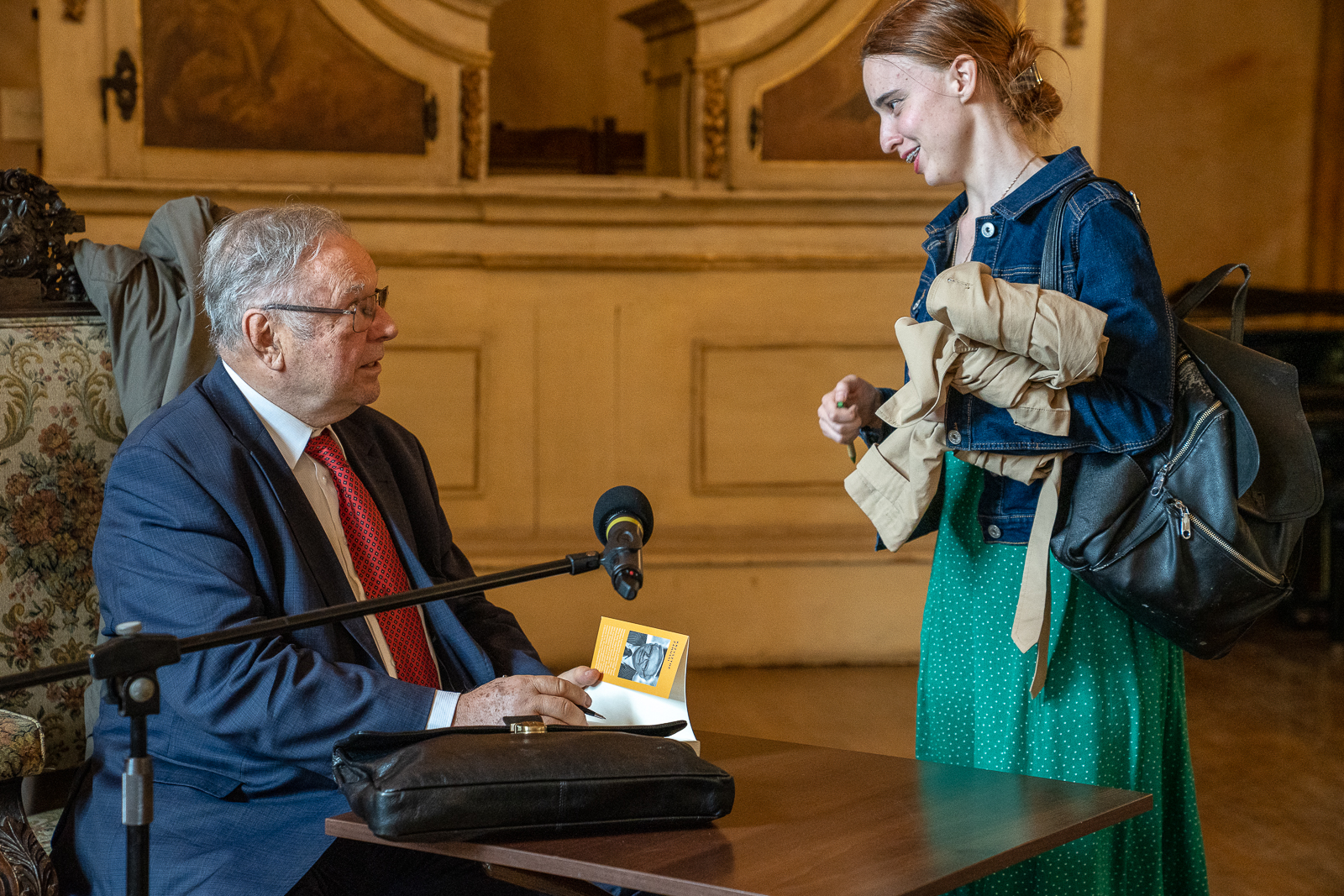 Młoda kobieta stoi przy stoliku, przy którym siedzi starszy mężczyzna z książką przy statywie z mikrofonem