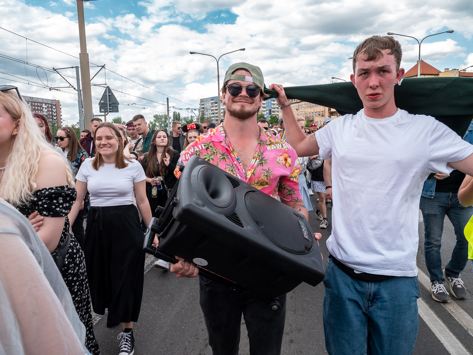 Ludzie idący w pochodzie juwenaliowym ulicami Wrocławia. Na pierwszym planie mężczyzna w różowej koszuli w kwiaty, trzyma w rękach wielki głośnik.