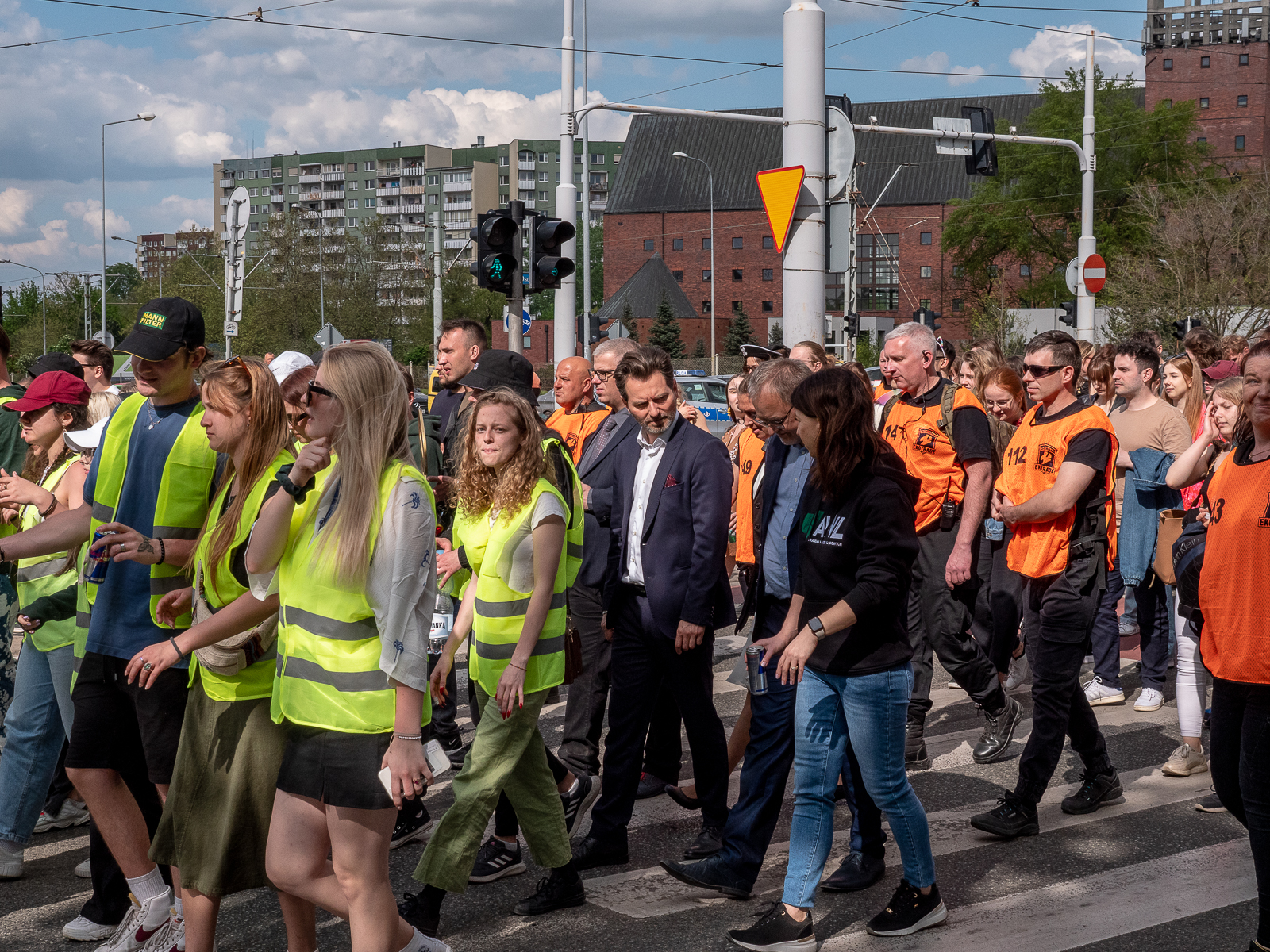 Grupa ludzi idąca razem ulicą miasta. Studenci w zielonych kamizelkach, ludzie w pomarańczowych kamizelkach. W środku męzczyna w garniturze.