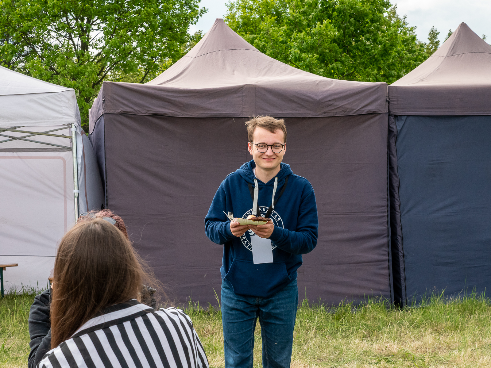 Uśmiechnięty student w okularach stoi na trawie, w tle fioletowe namioty, przed nim na pierwszym planie odwrócona tyłem studentka w koszuli w biało-czarne pasy.