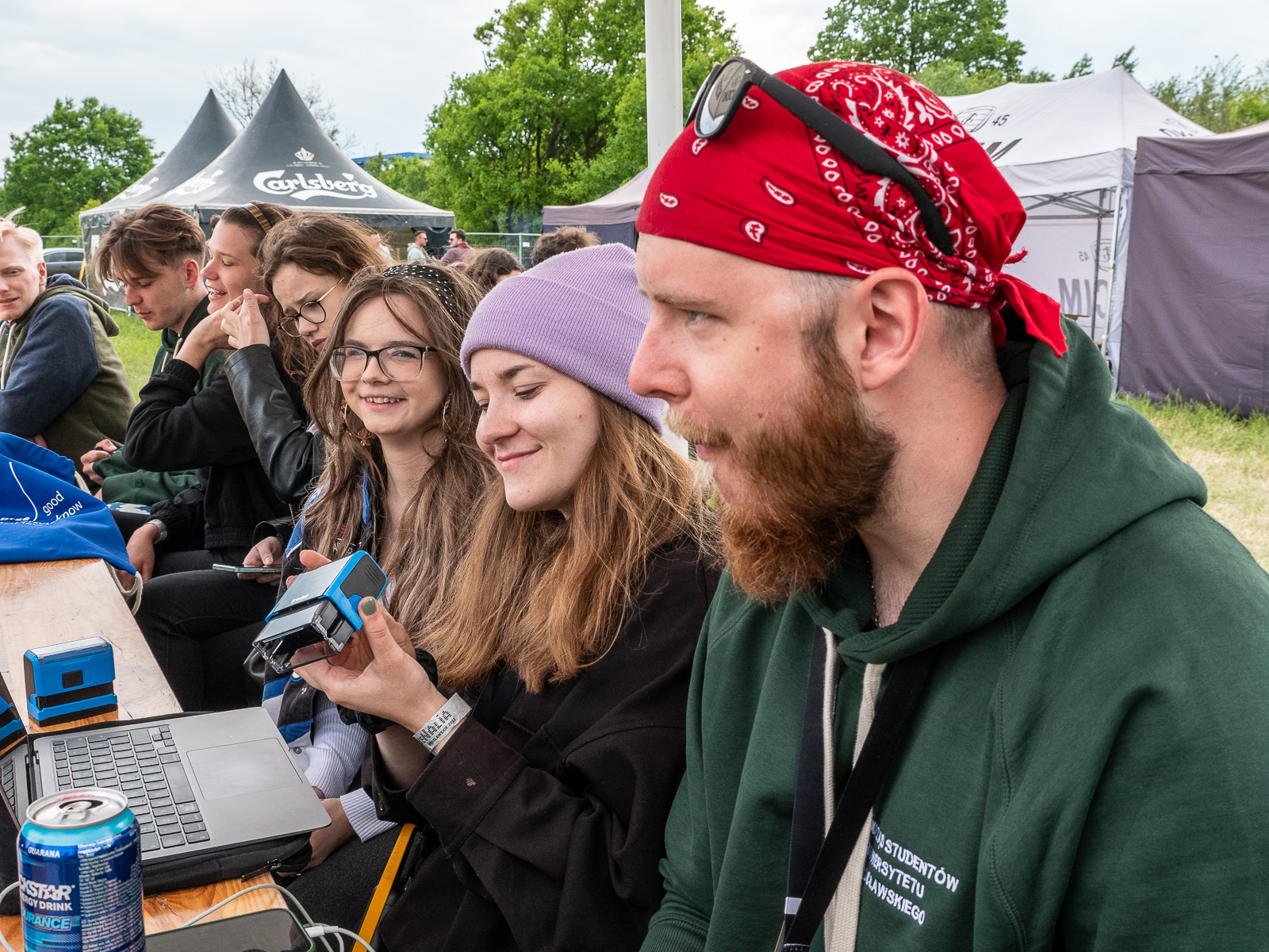 Grupa studentów siedząca przy stole. Widać laptop.Jedna studentka ma w ręku pieczątkę. Na pierwszym planie student w czerwonej chustce na głowie.