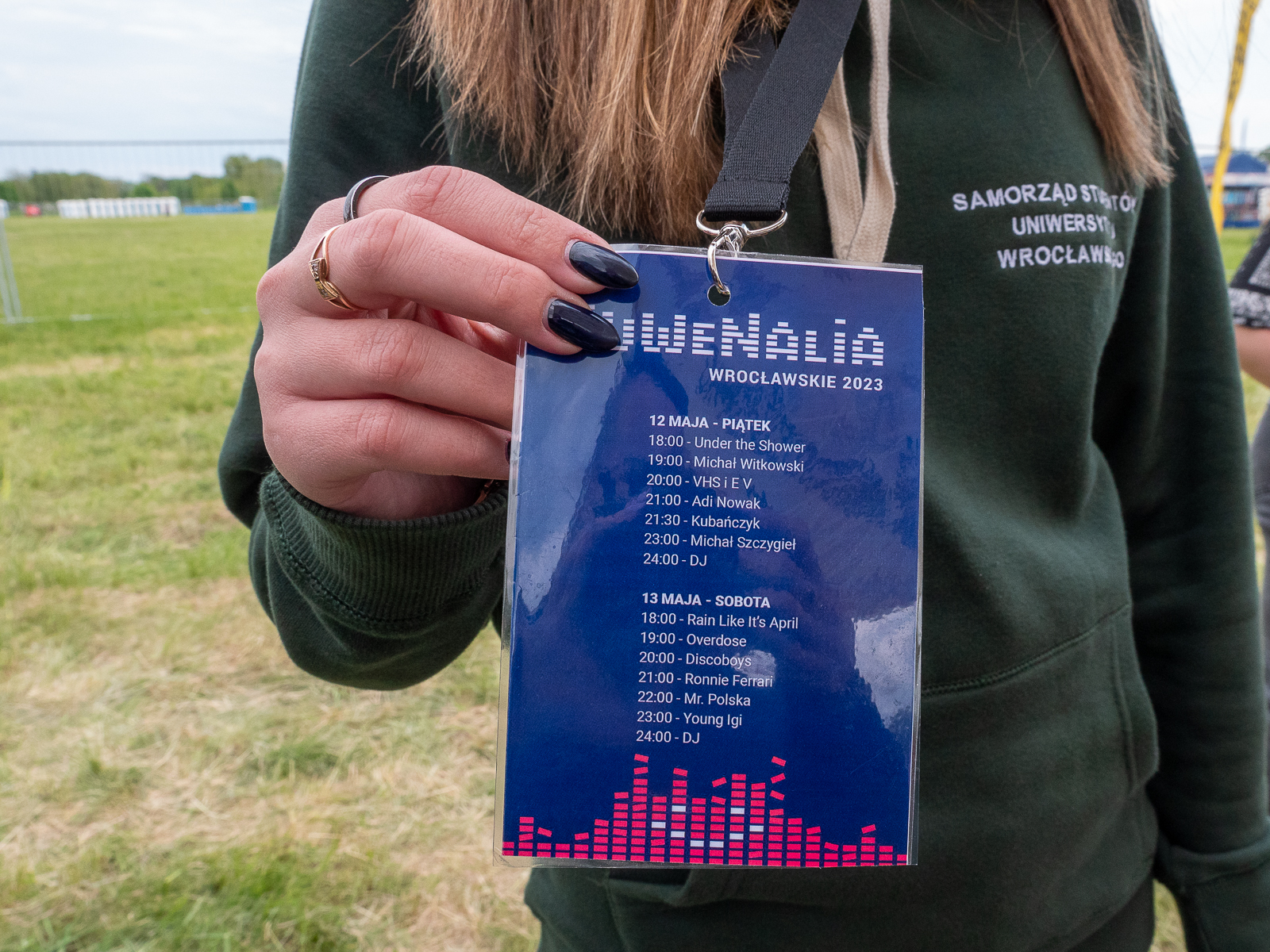 Plakietka z napisem Juwenalia Wrocławskie 2023 i programem koncertów, która wisi na szyi studentki w zielonej bluzie z napisem Samorząd Studentów Uniwersytetu Wrocławskiego. Studentka trzyma plakietkę dwoma palcami.