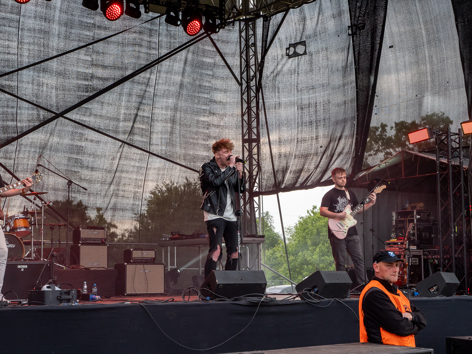 Mężczyzna na scenie koncertowej śpiewa do mikrofonu, po jego prawej stronie mężczyzna grający na gitarze. Pod sceną plecamid o niej mężczyzna - pracownik ochrony - w pomarańczowej kamizelce.